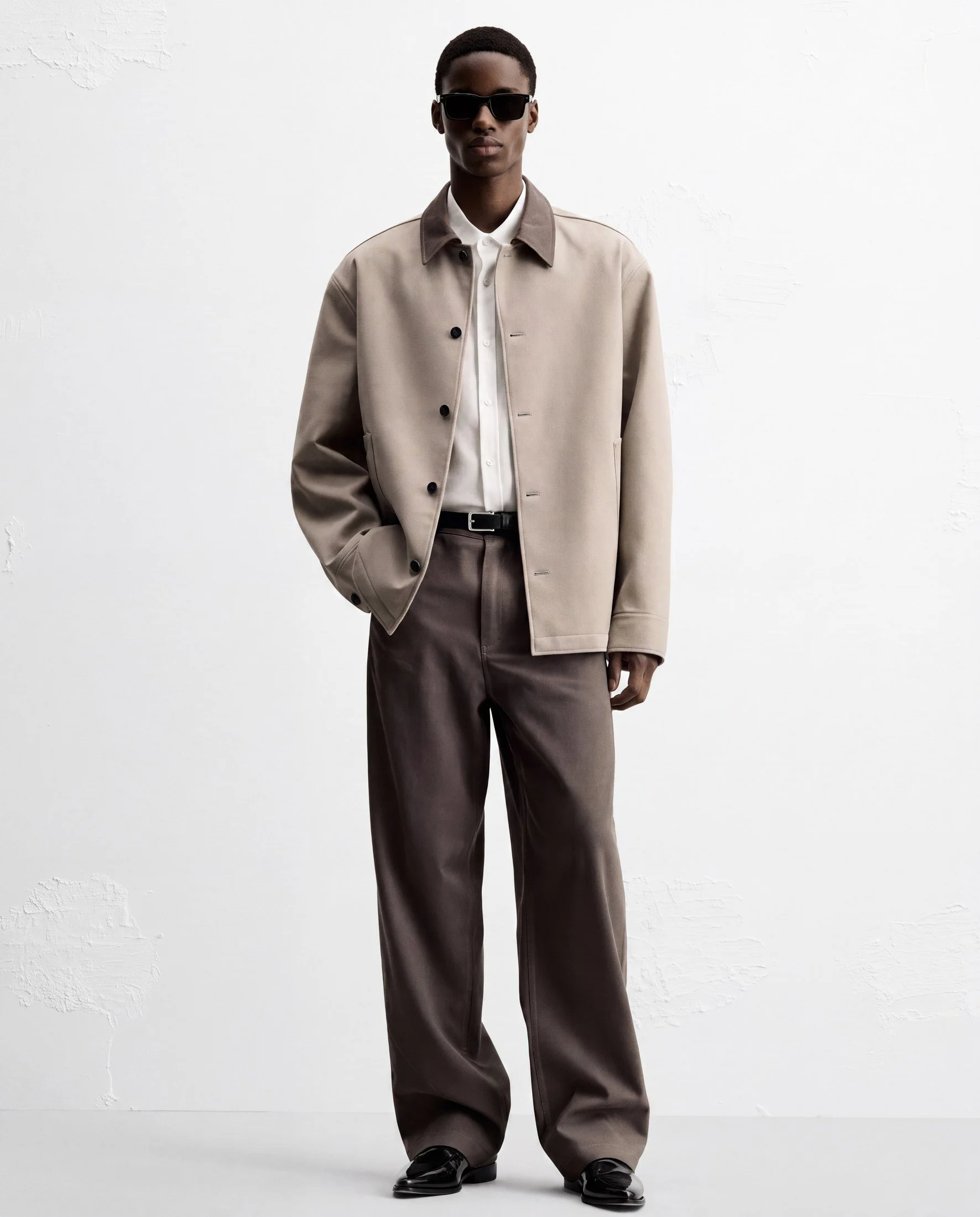 A tall person with short hair wearing black sunglasses, a beige jacket with a darker collar, a white button-up shirt, loose brown pants, and black shoes, standing against a plain white wall.