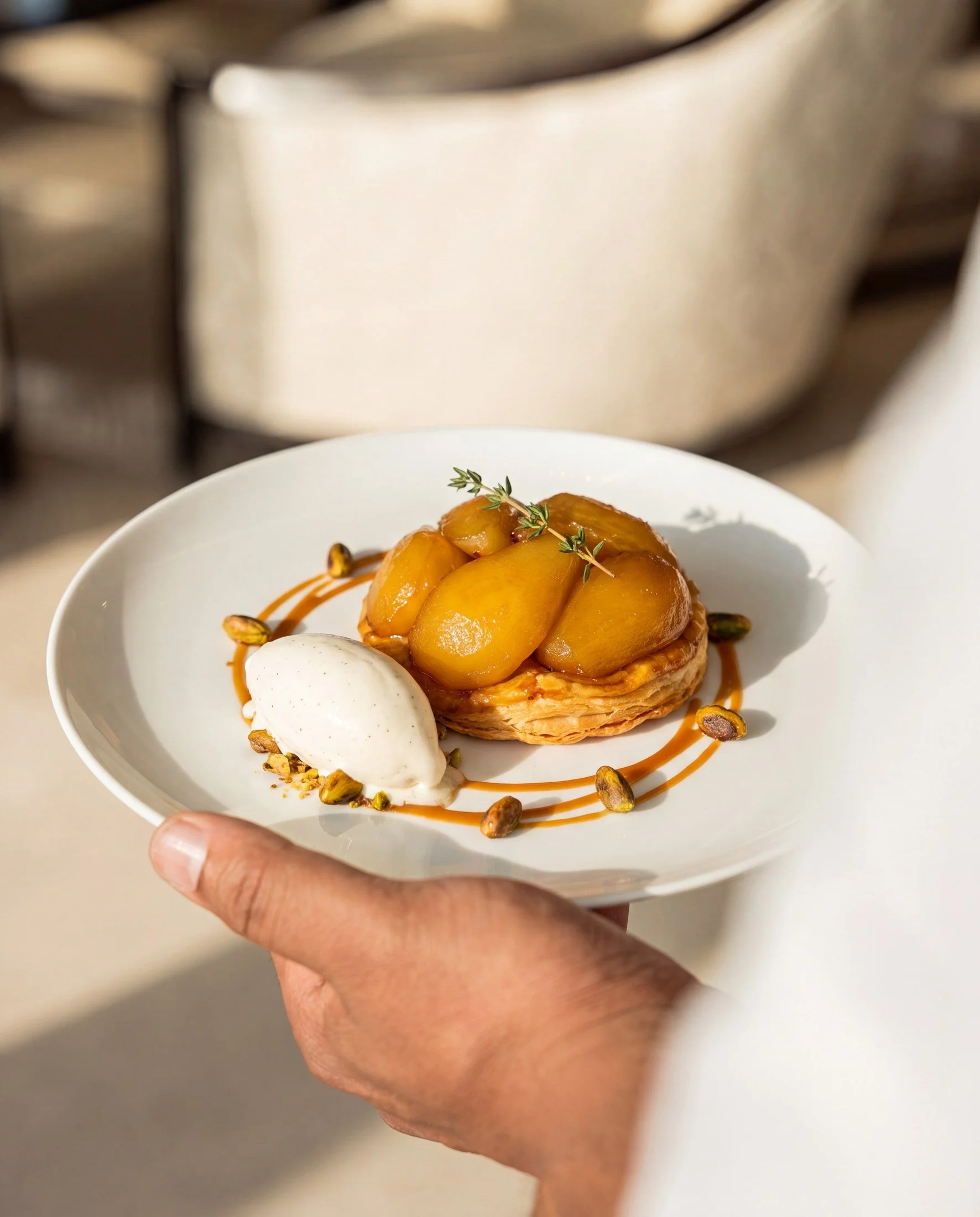 A hand holding a white plate with a dessert featuring peach slices, a scoop of ice cream or whipped cream, caramel sauce, and garnished with small green herbs and pistachios.