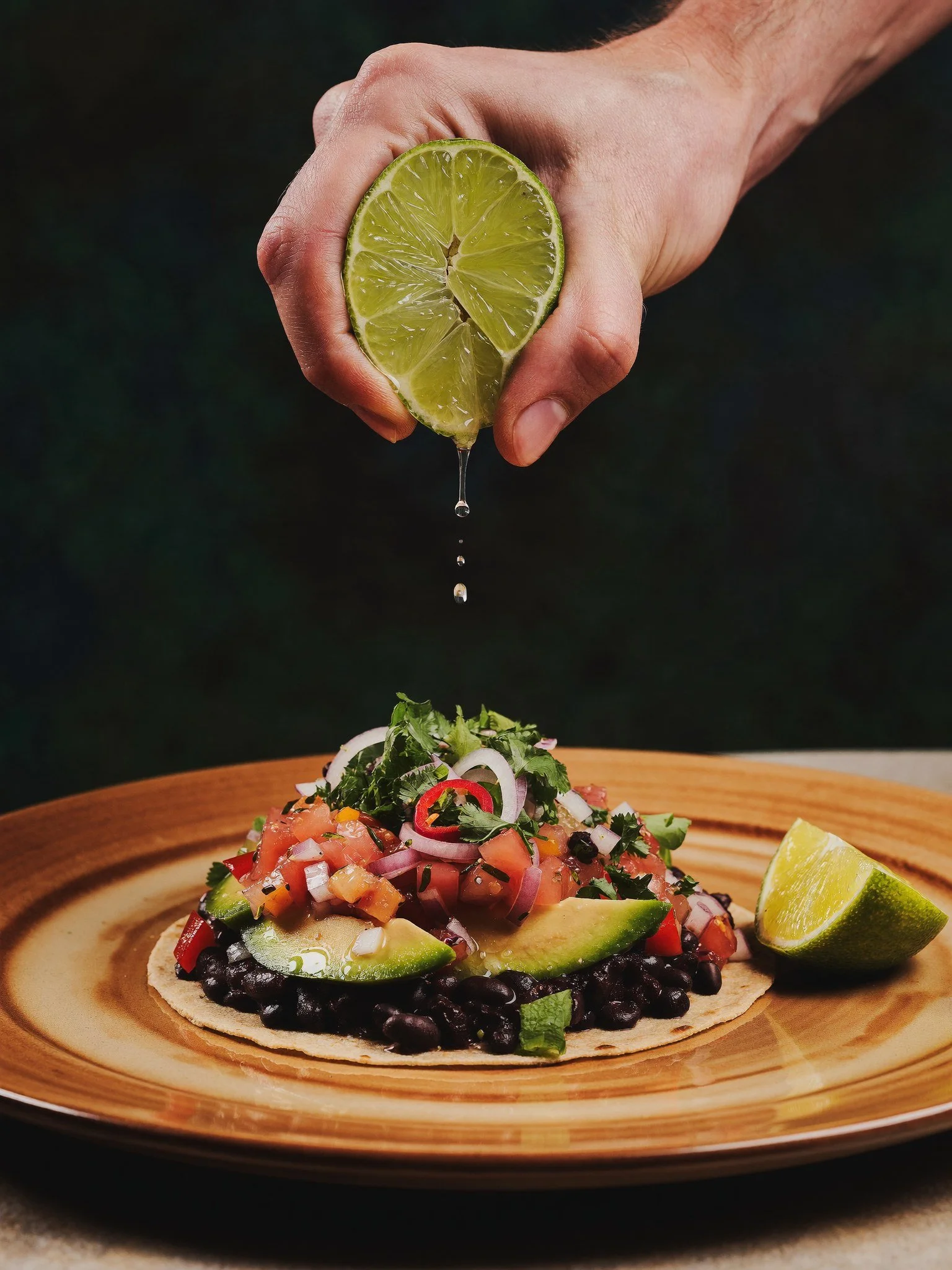 A hand squeezing a lime over a plate of food, which appears to be a taco topped with diced vegetables, chopped cilantro, onions, and sliced red chili peppers, with lime wedges on the side.