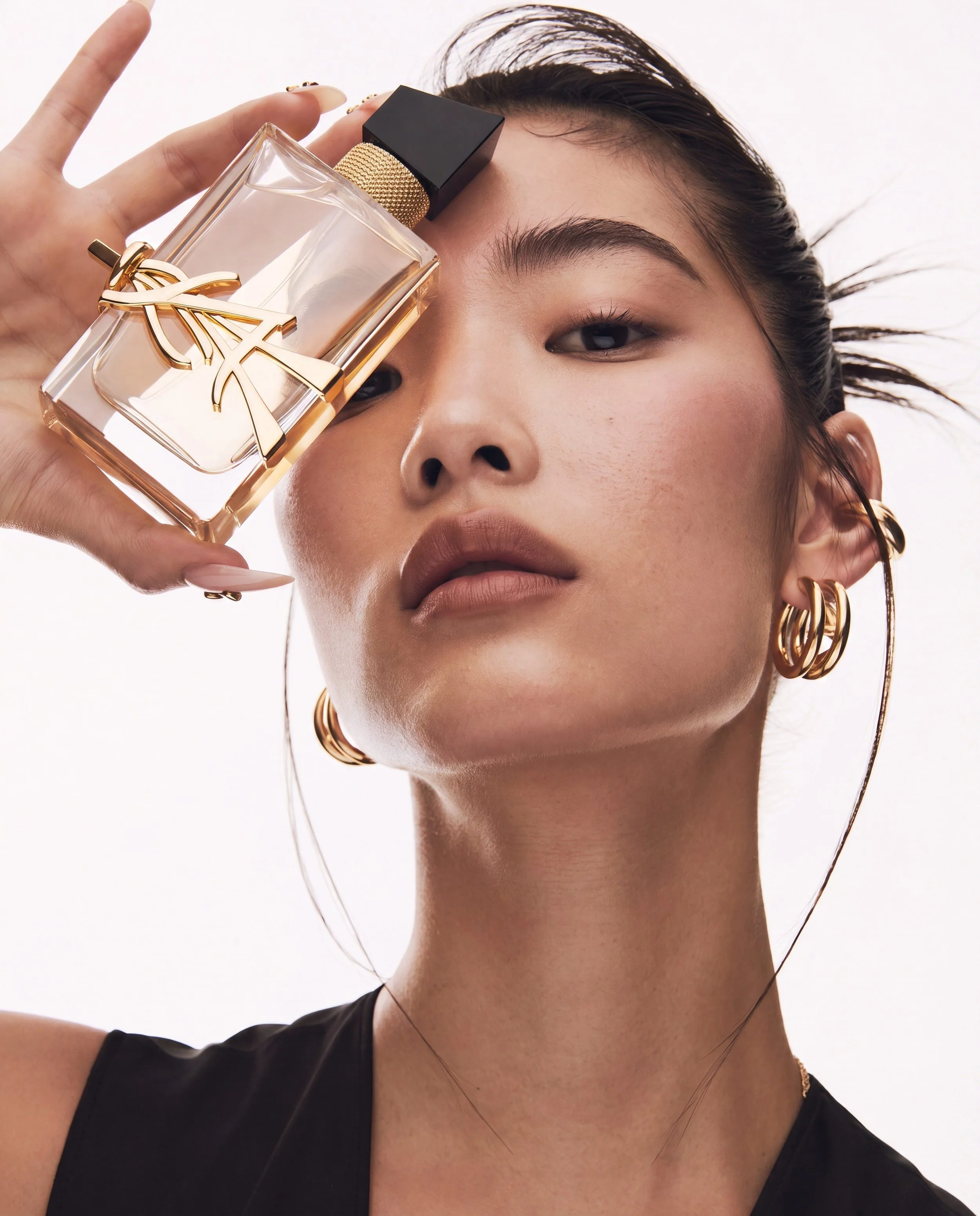 A woman holds a bottle of Yves Saint Laurent perfume close to her face. She has dark hair, gold hoop earrings, and wears a black top and necklace.