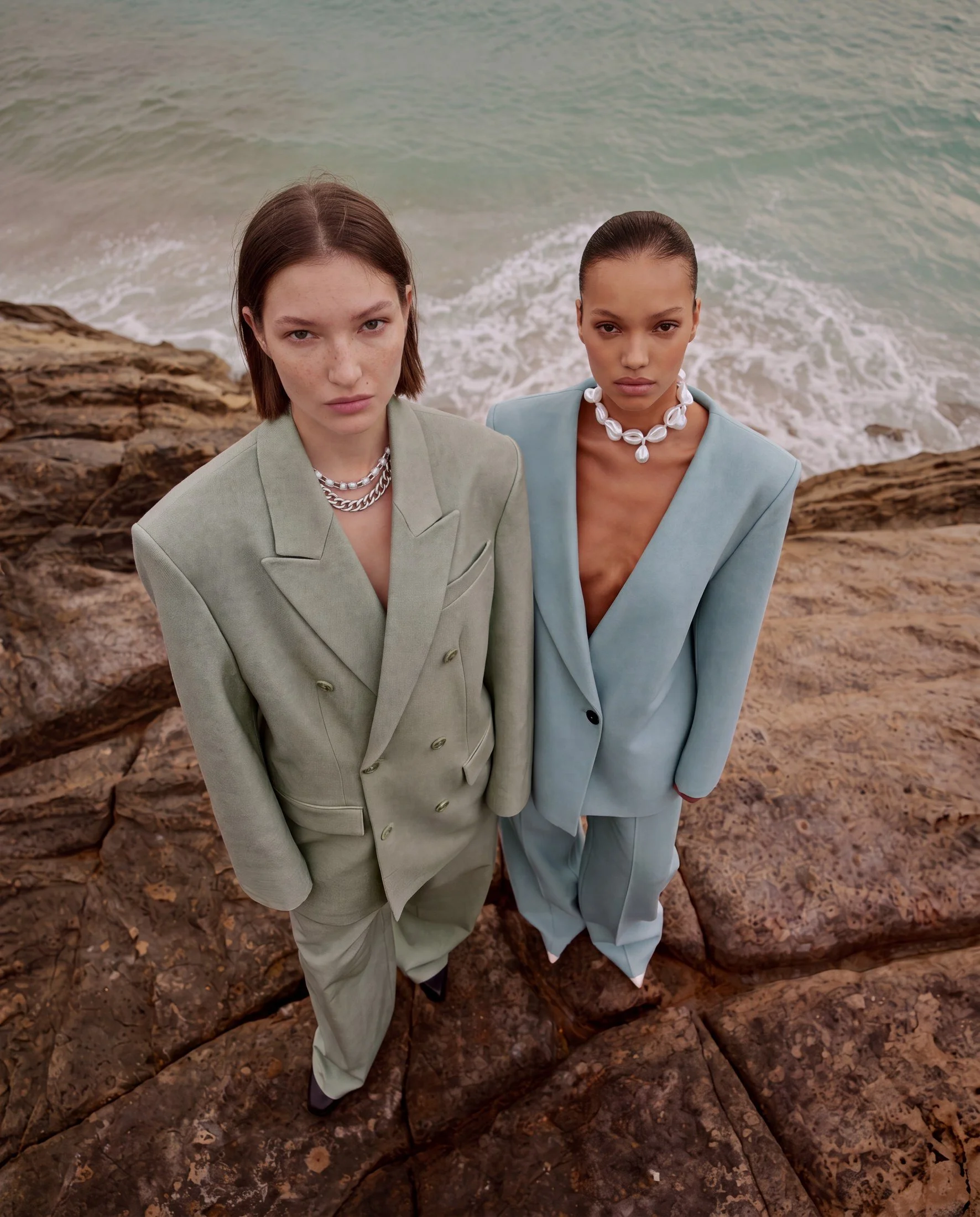 Two women standing on rocks at the beach, looking into the camera with serious expressions, dressed in pastel suits with jewelry, ocean background.