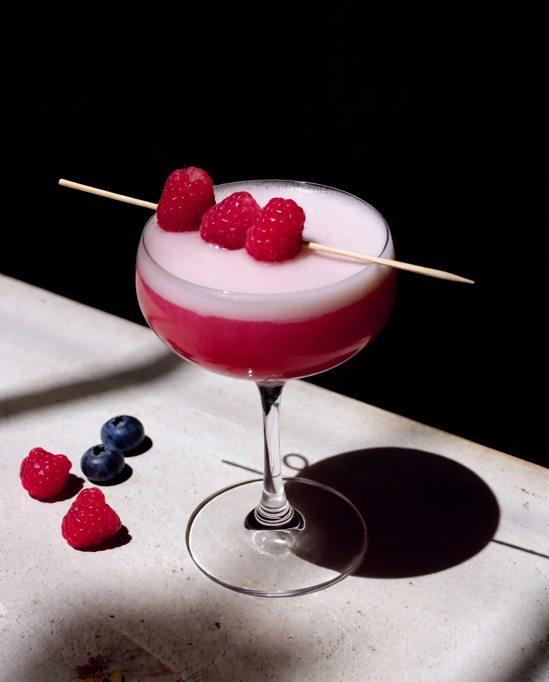 A pink cocktail in a martini glass garnished with three raspberries, with additional raspberries and blueberries spilled on a white surface nearby, against a black background.