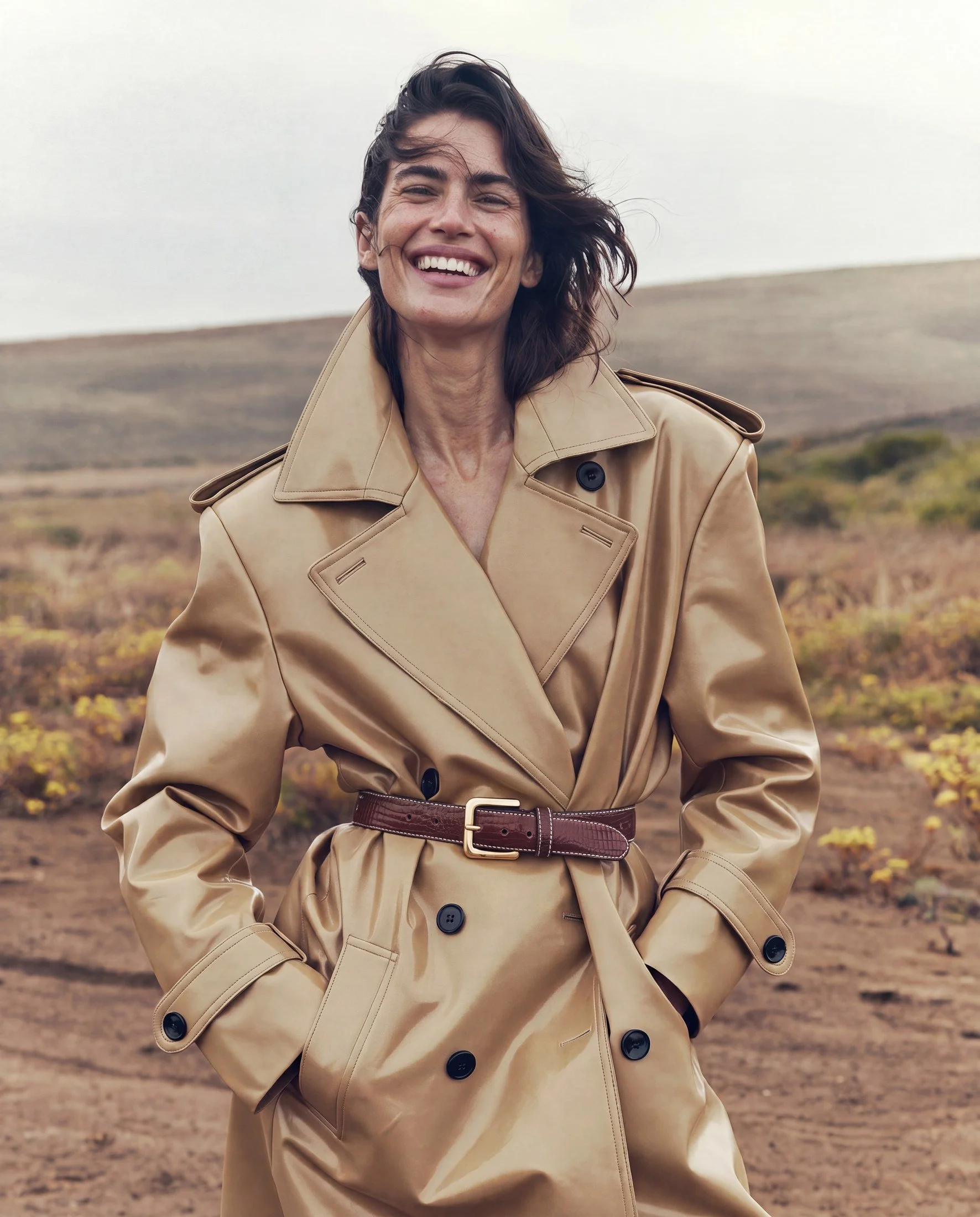 A woman smiling outdoors in a beige trench coat with a brown belt, standing in a field with low hills and sparse vegetation under a cloudy sky.