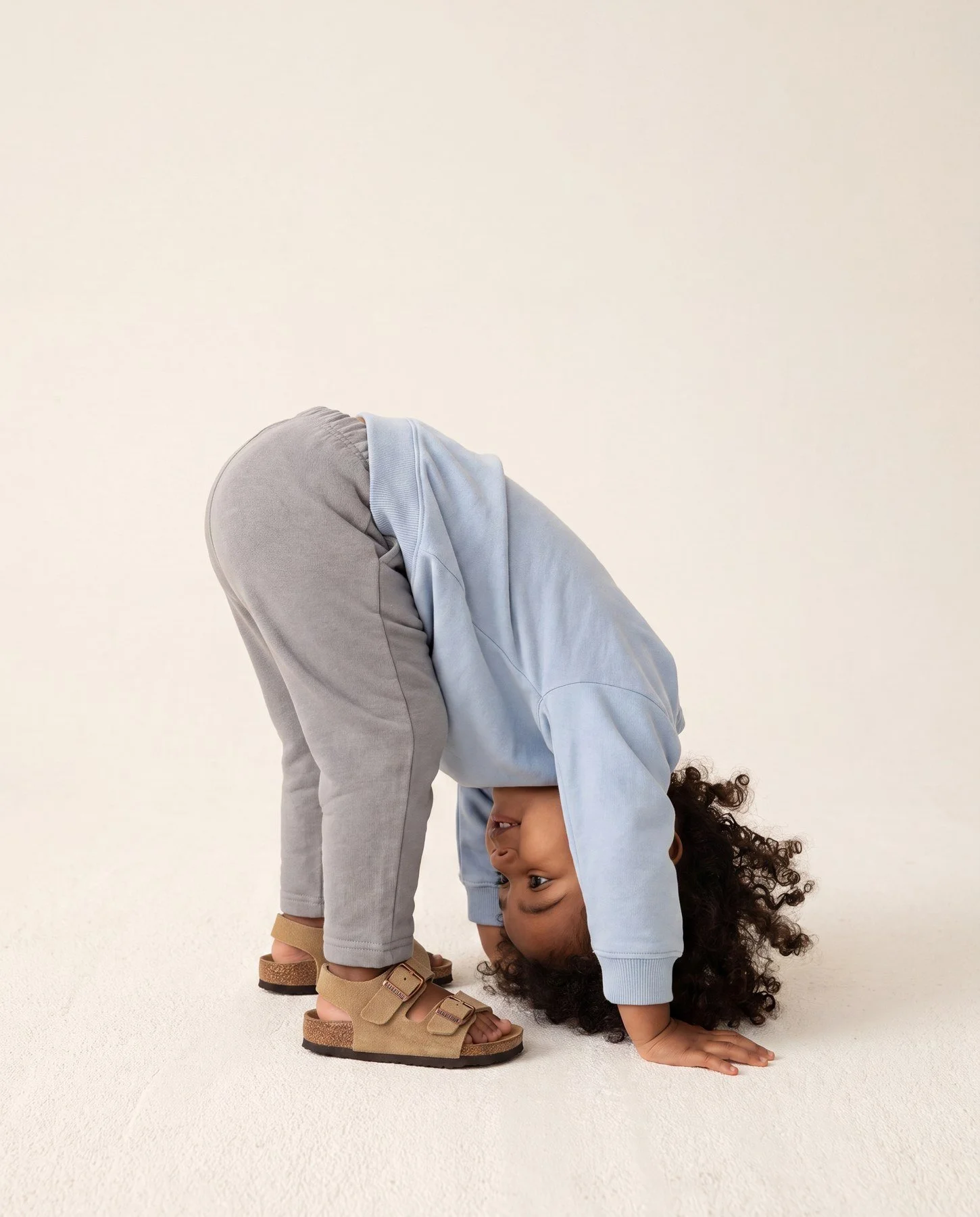 A child in a light blue sweatshirt and gray pants is bending over with hands and feet on the ground, head upside down, with curly hair hanging down, on a white background
