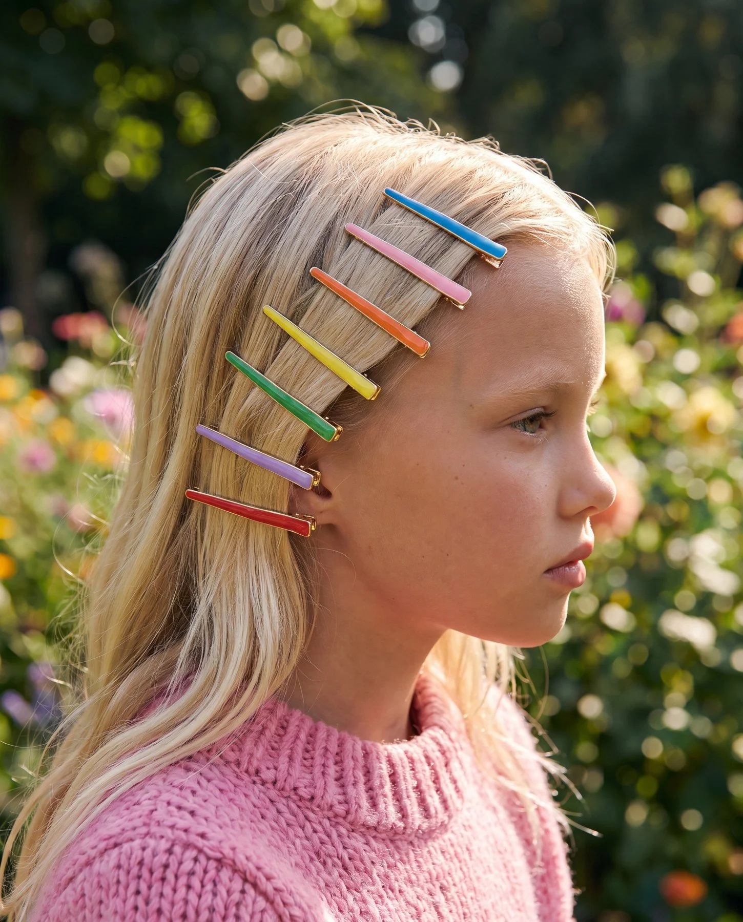 Profile of a young woman with blonde hair wearing a pink sweater, with colorful hair clips in her hair, standing outdoors with blurred greenery and flowers in the background.