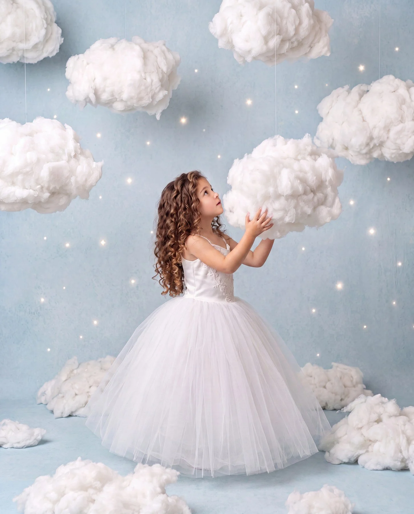 A young girl dressed as a fairy or princess in a white gown with a full skirt, holding a fluffy cloud decoration in a sky-themed setting with hanging clouds and star-like lights.