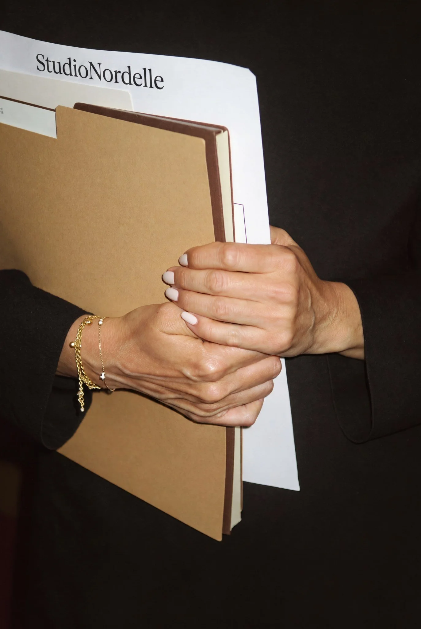 Person holding a thick brown folder and a white sheet of paper, wearing a black long-sleeve shirt and gold jewelry on their wrist.