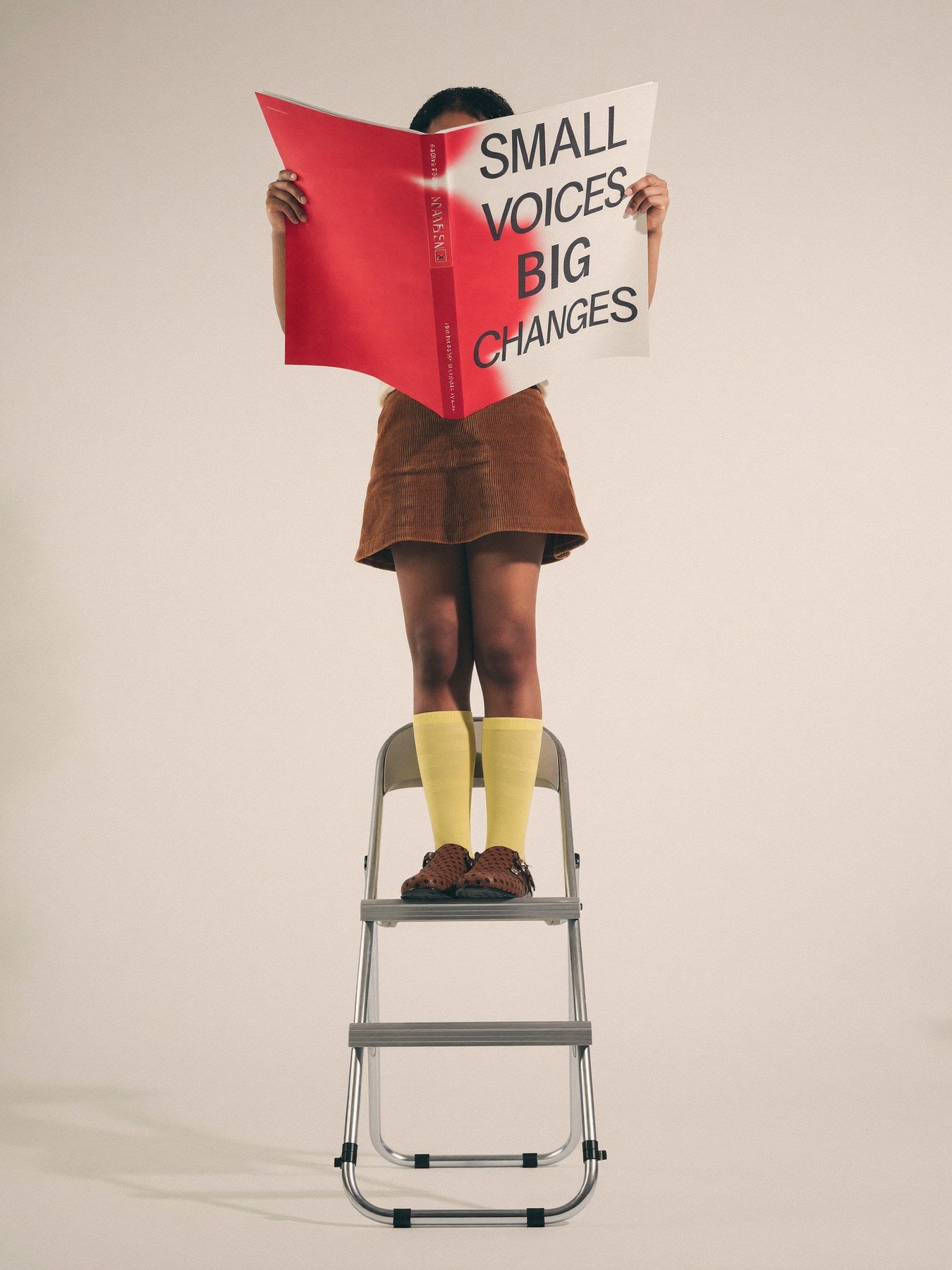 Person standing on a step ladder, reading a large book that covers their face, with a plain white background.