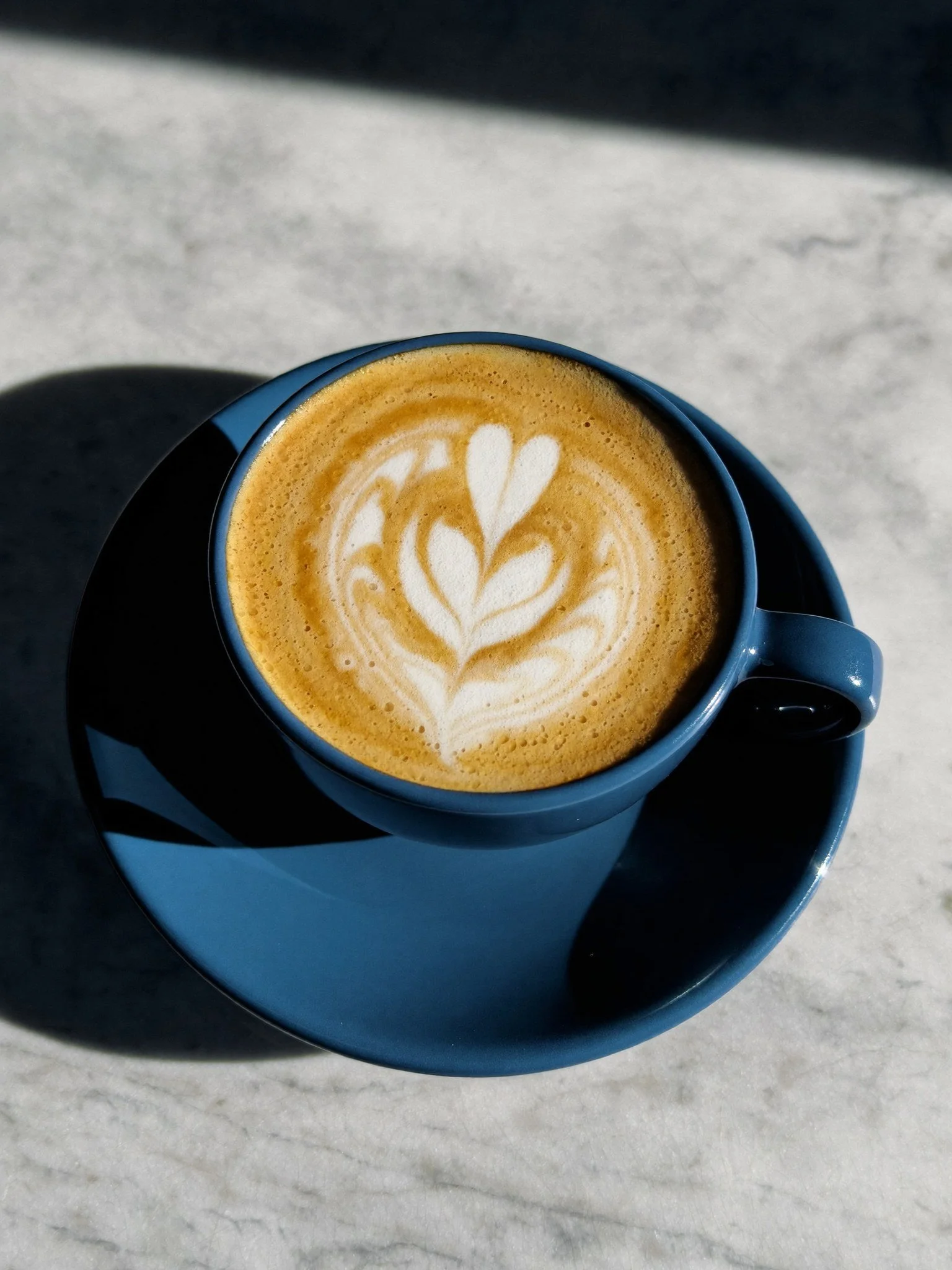 A blue ceramic cup filled with latte art coffee, placed on a matching blue saucer on a white marble surface.