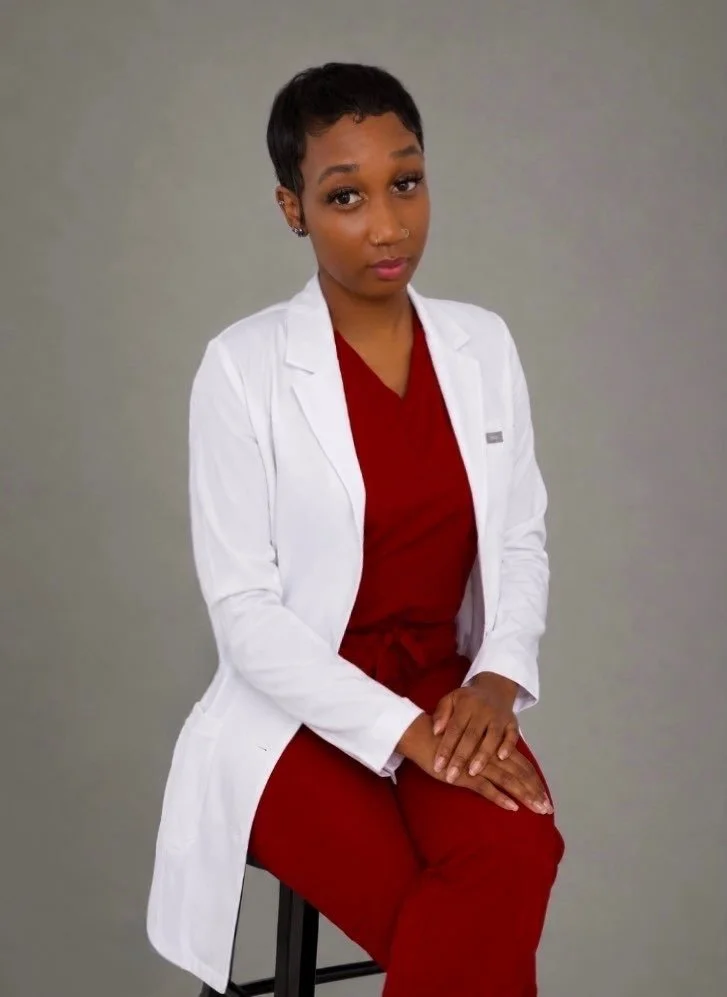 A woman with short black hair wearing a white lab coat over maroon scrubs, sitting on a stool against a plain gray background.
