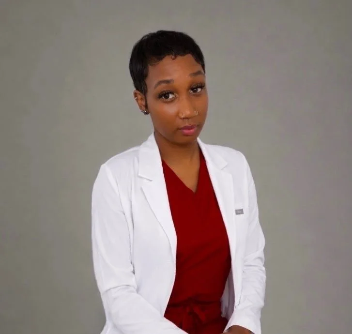 Portrait of a young woman with short hair wearing a white lab coat and a red scrub top, sitting against a neutral background.
