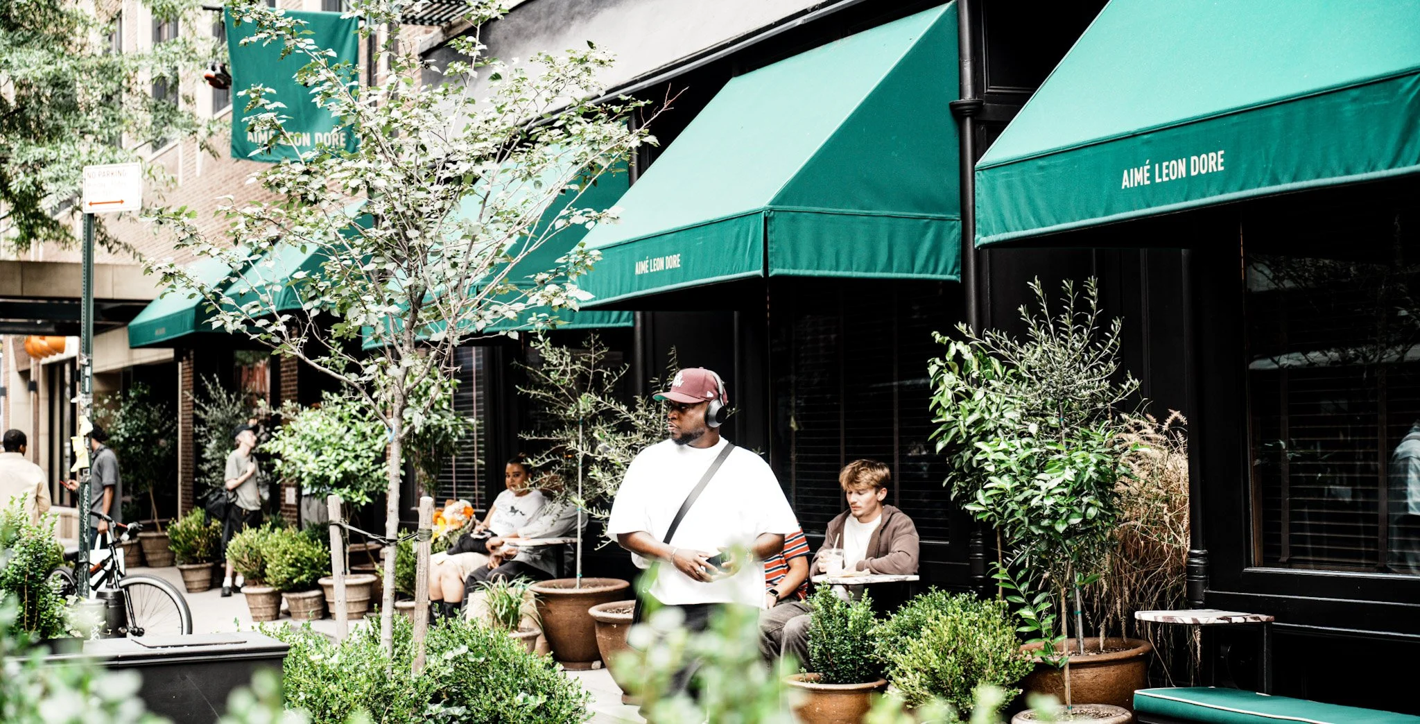People sitting and standing outside a storefront with green awnings labeled 'Aimé Leon Dore,' surrounded by potted plants and trees, on a city street.