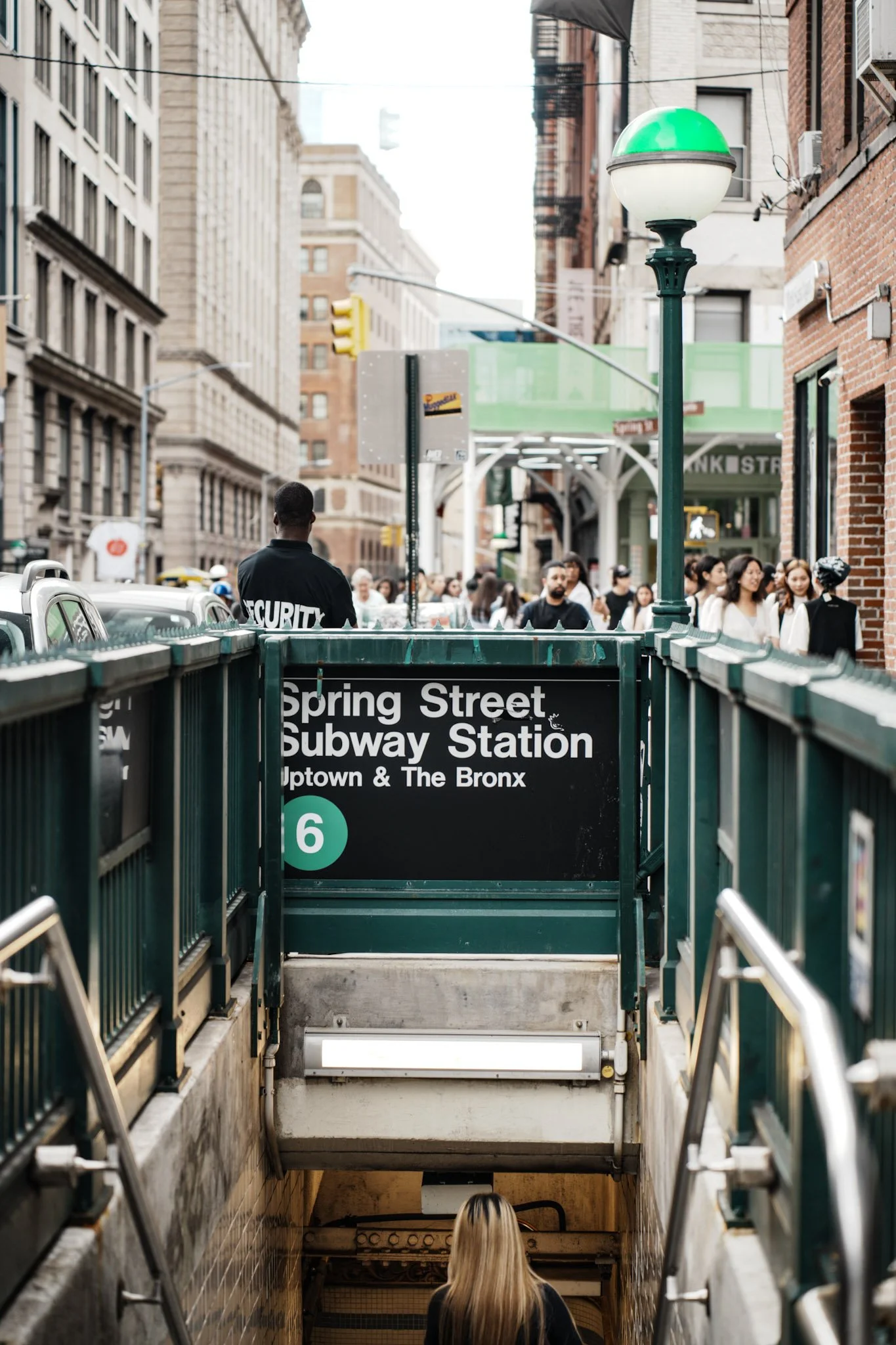Entrance to Spring Street Subway Station with a woman walking down the stairs, surrounding buildings, and a green and white street lamp.
