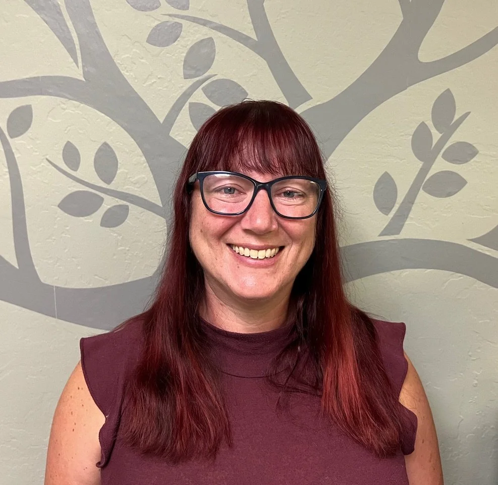 Woman with long red hair and glasses smiling in front of a wall with a tree and leaf pattern.