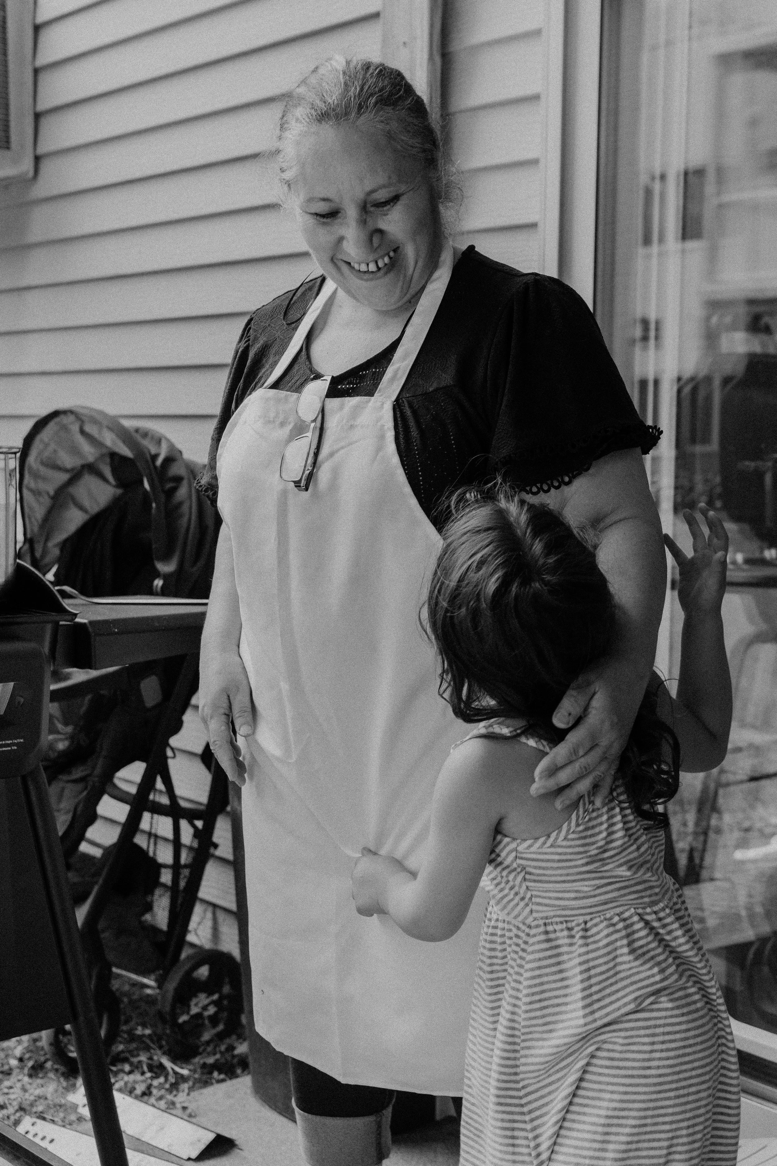 A smiling woman with glasses hanging from her neck stands next to a young girl who is hugging her. They are outside, near a house with horizontal siding, and there are various items including a stroller in the background.