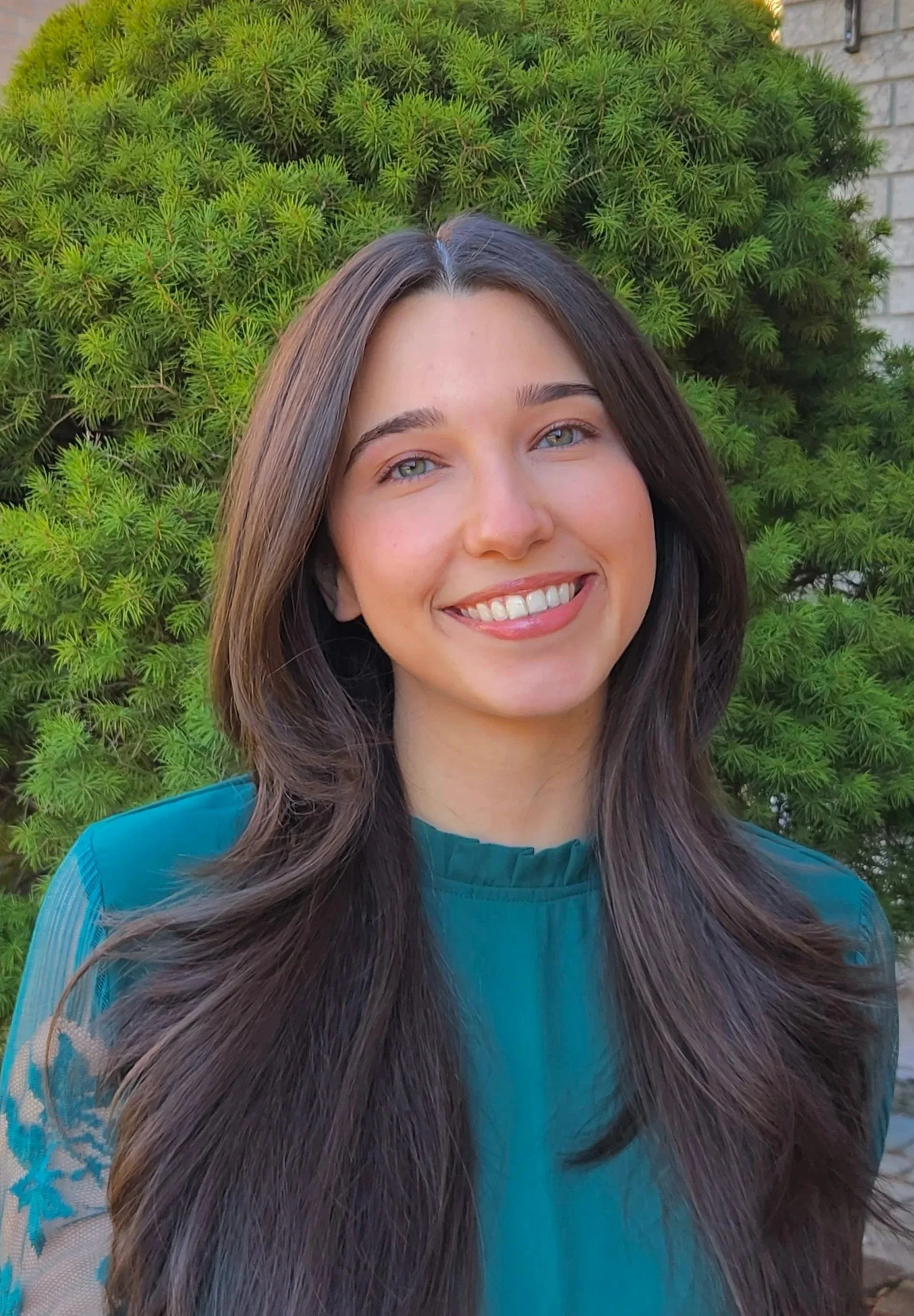 A young woman with long brown hair, blue eyes, and a bright smile, wearing a teal top with lace details on the sleeves. She is standing outdoors in front of a lush green bush and a brick wall.