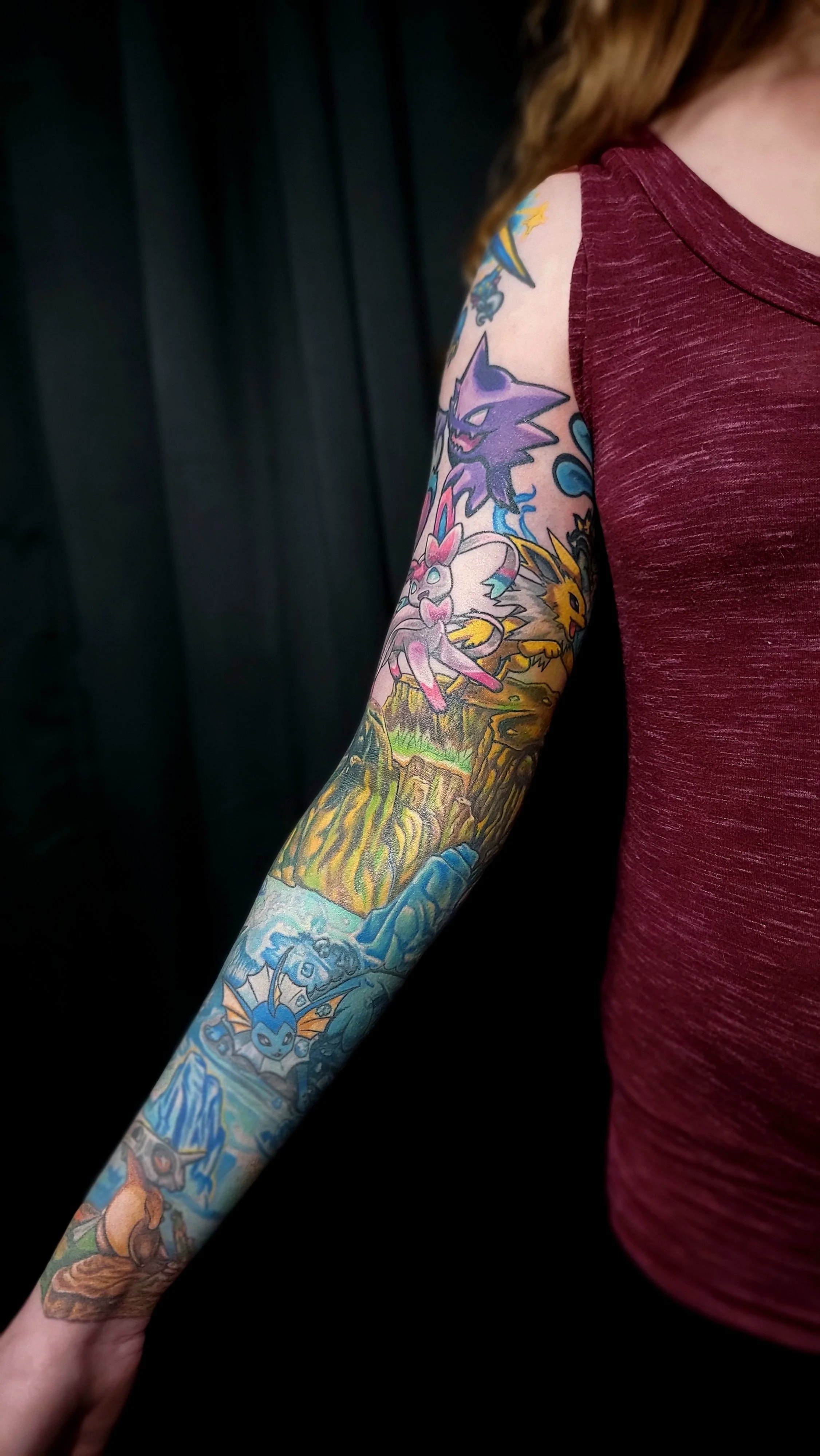 Close-up of a person's arm with a colorful tattoo sleeve featuring various Pokémon characters, partially covered by a maroon T-shirt, against a black background.