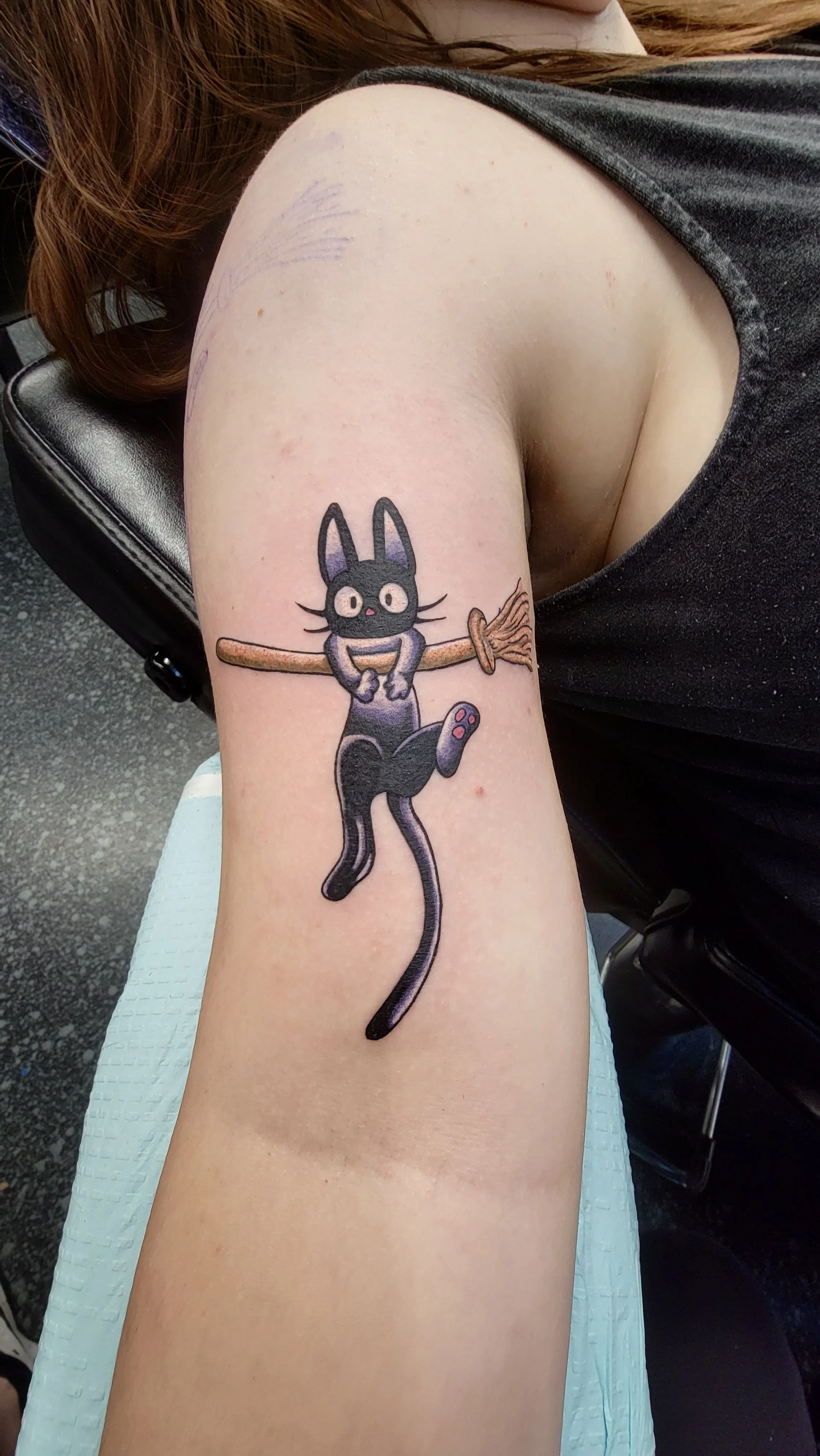 Tattoo of a black cartoonish cat with big eyes and a pink nose, holding a broomstick with a string, on a person's upper arm.