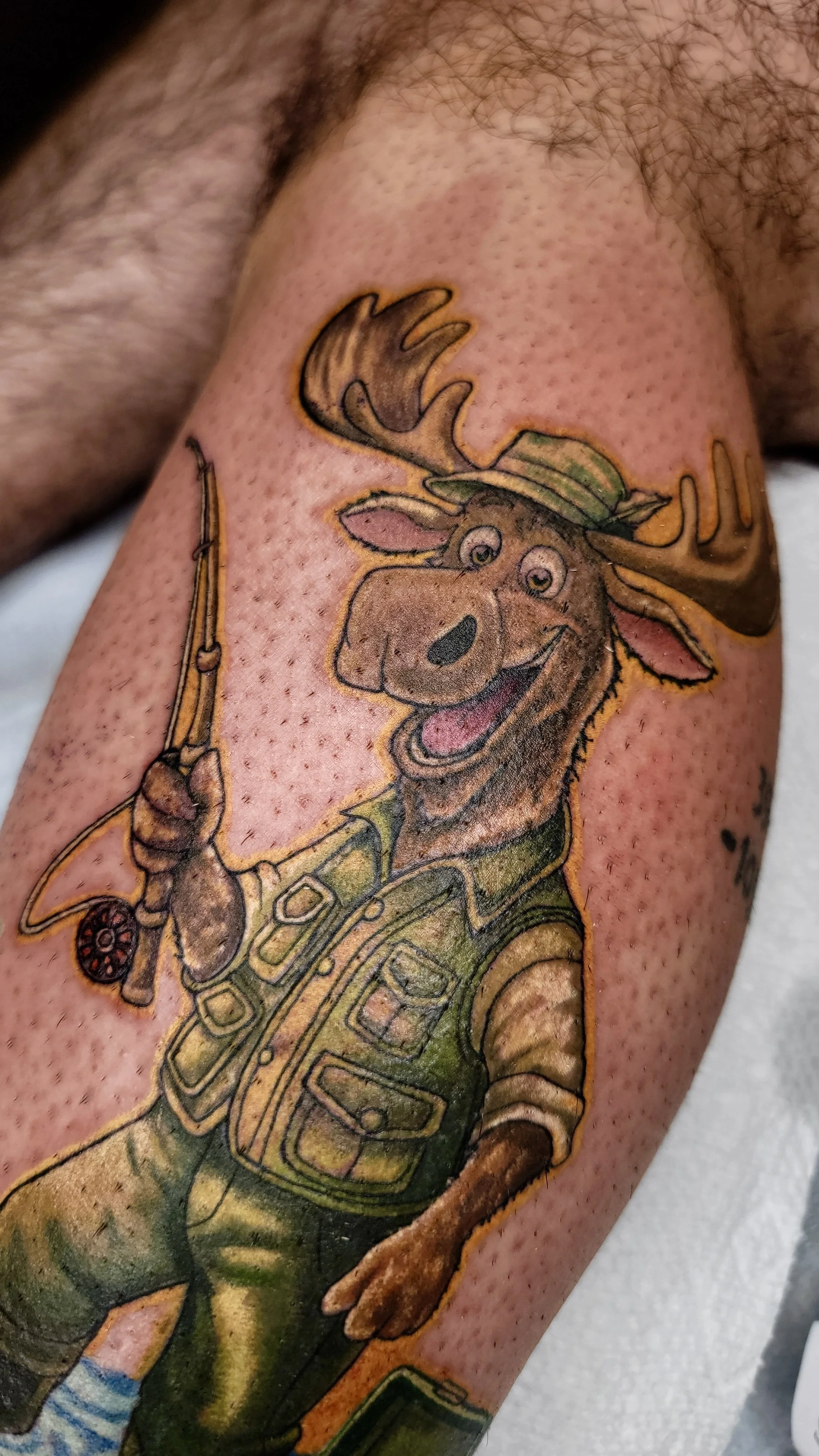 Tattoo of a cartoon moose holding a fishing rod, dressed in military or scout uniform, with antlers and a happy expression.