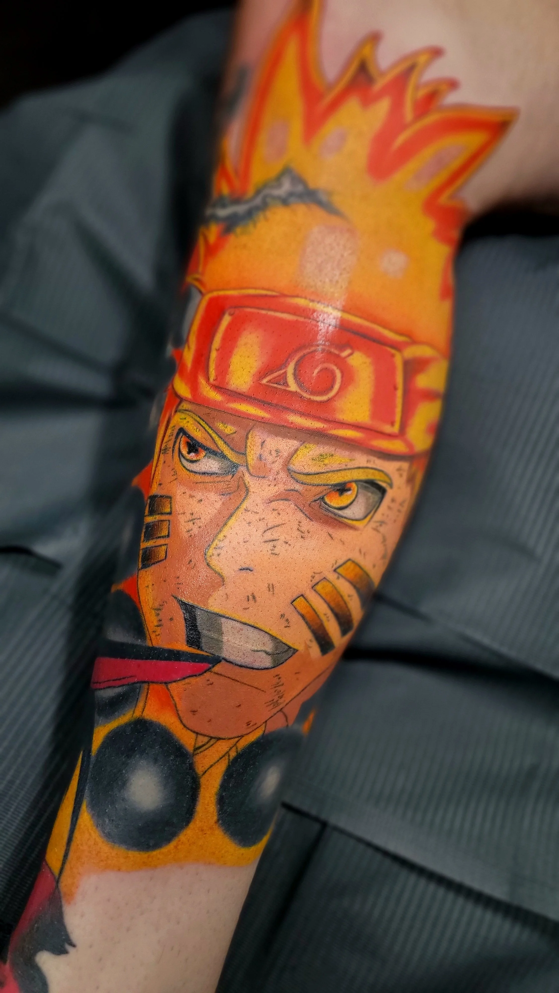 Tattoo of Naruto Uzumaki's face from the anime Naruto, with a background of orange and fiery design, on a person's arm.