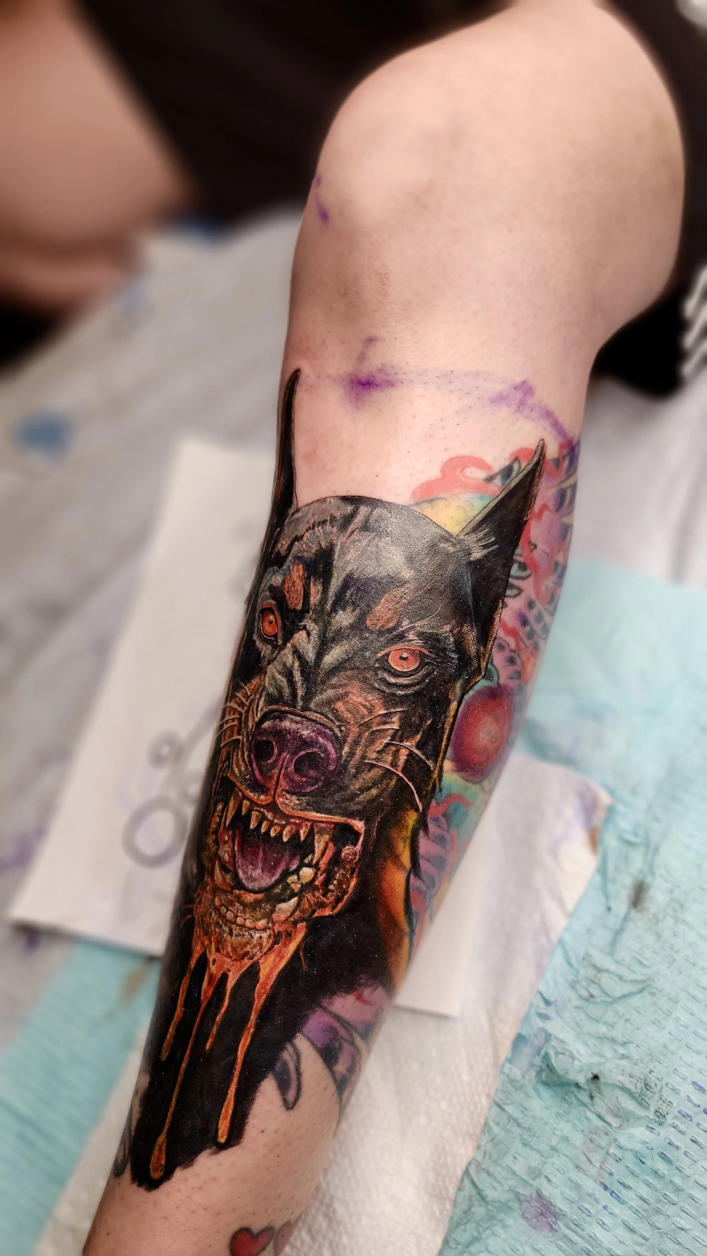 A person's arm with a colorful tattoo of a snarling wolf with red eyes and dripping orange and yellow accents, on a bright background with purple, pink, and green elements.