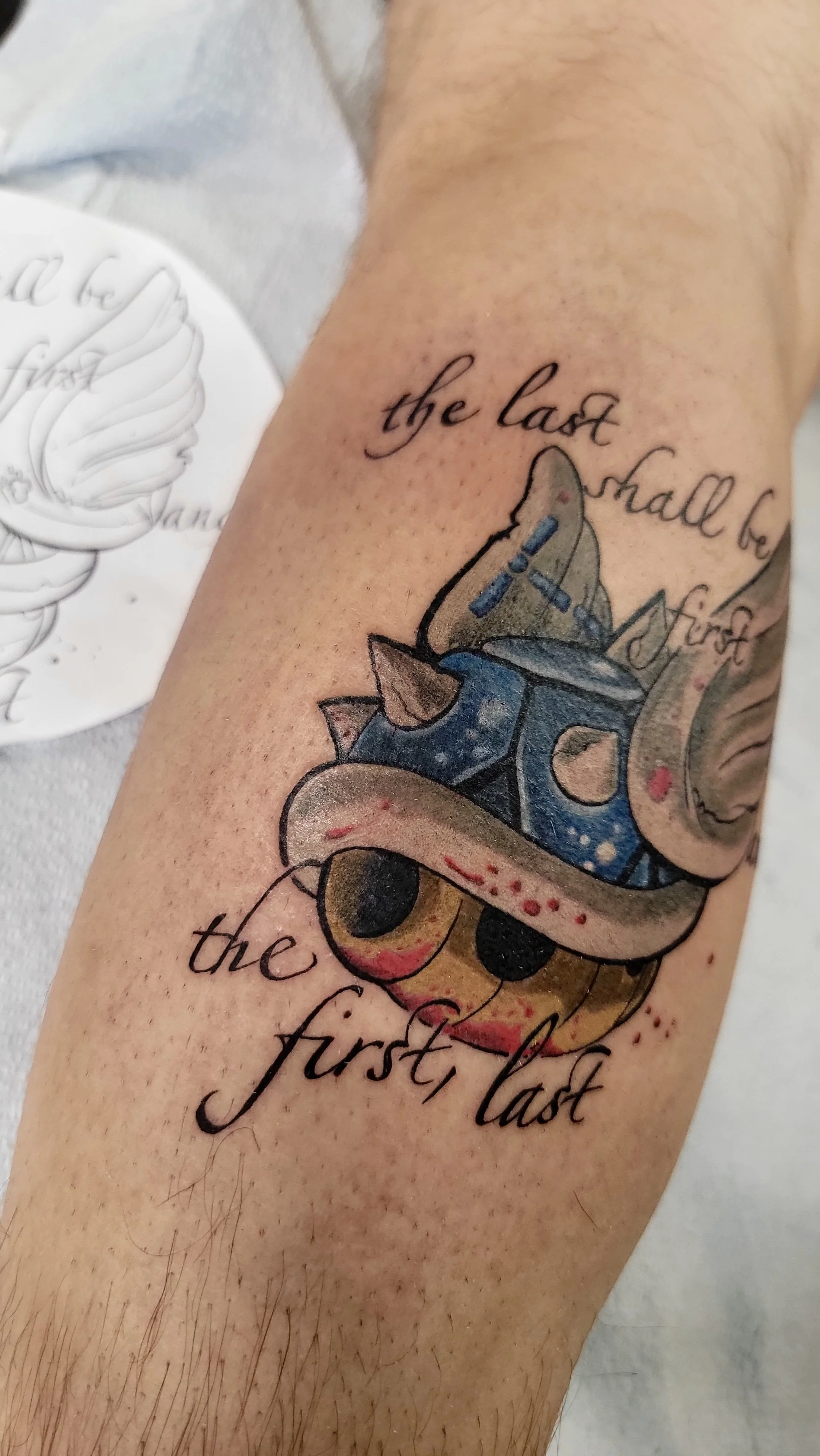 Close-up of a tattoo on a person's arm with the words "the last will be the first" and a colorful cartoonish dog wearing a crown.