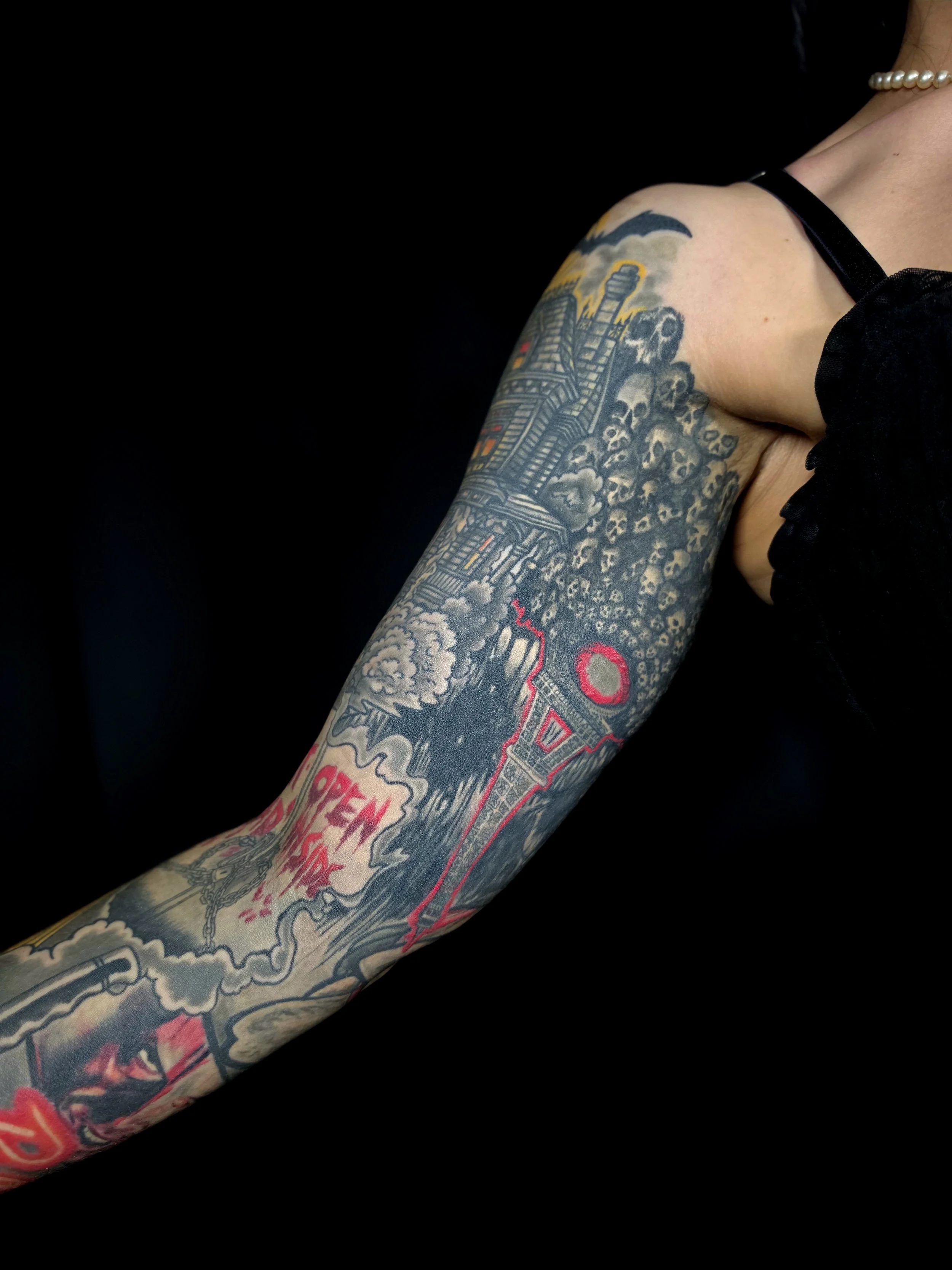 Close-up of a person's arm with a colorful tattoo featuring buildings, smoke, skulls, and bold red lettering.