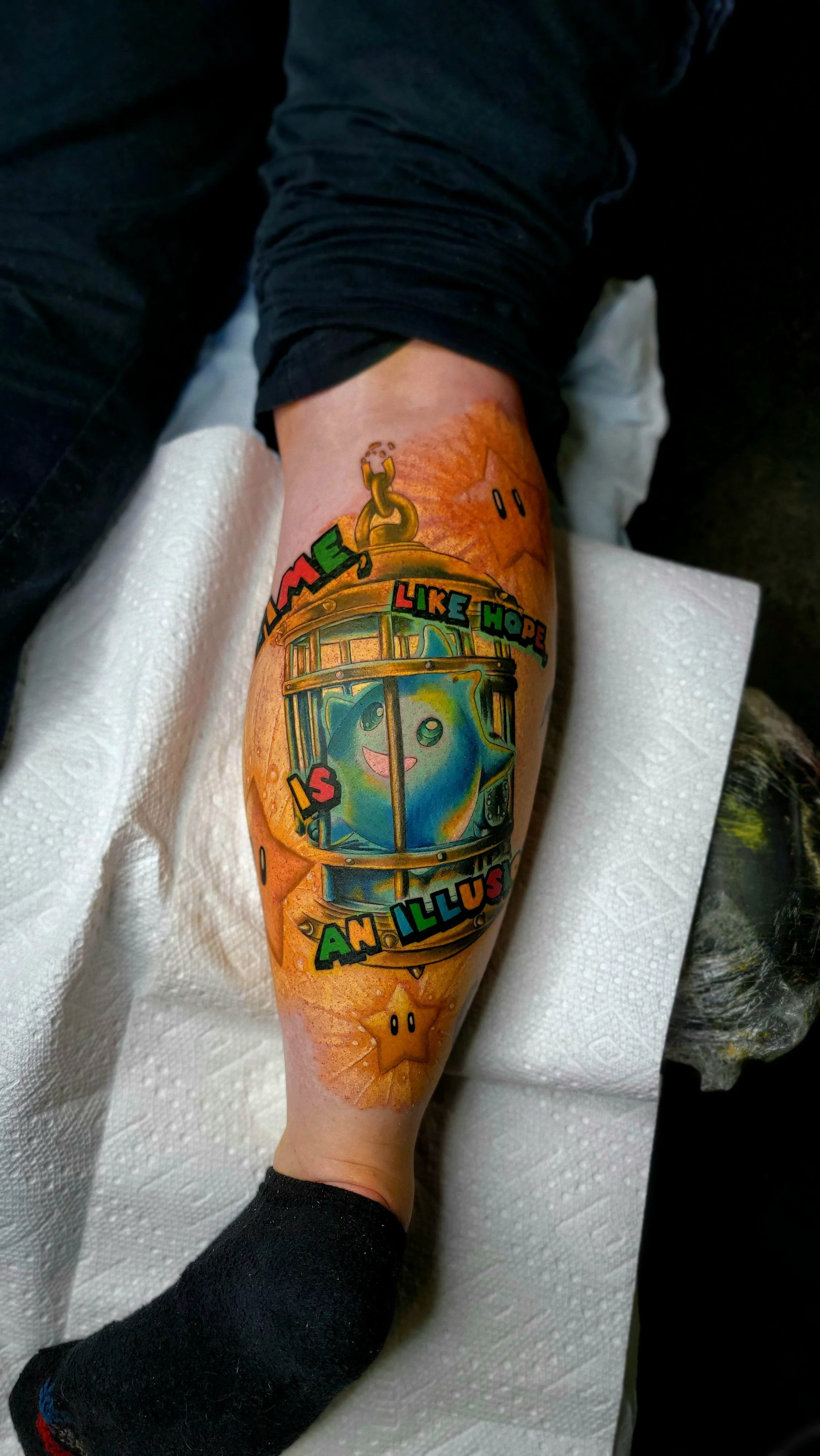 Colorful tattoo on person's arm featuring a cartoonish train with a smiling star character inside, surrounded by colorful text that reads 'LIKE HOPE IS AN ILLUSION' and other small stars and designs.