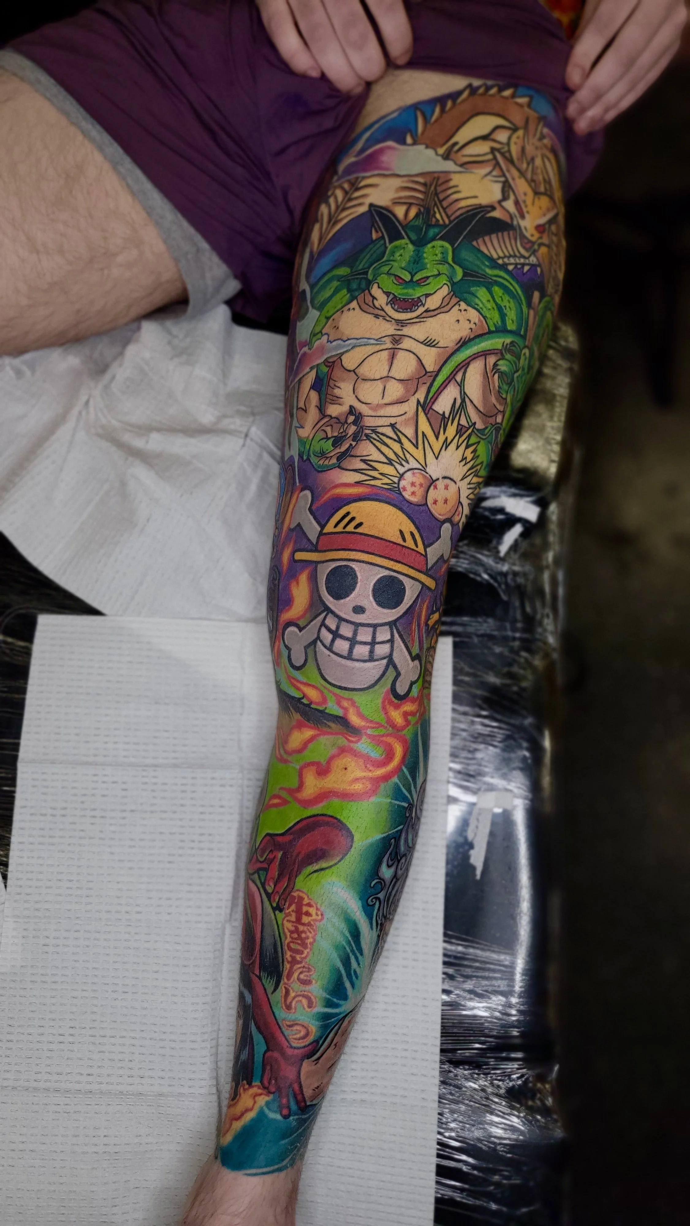 Colorful full sleeve tattoo featuring a green dragon, a skull with a straw hat, a fiery dragon head, and various vibrant flames and clouds.