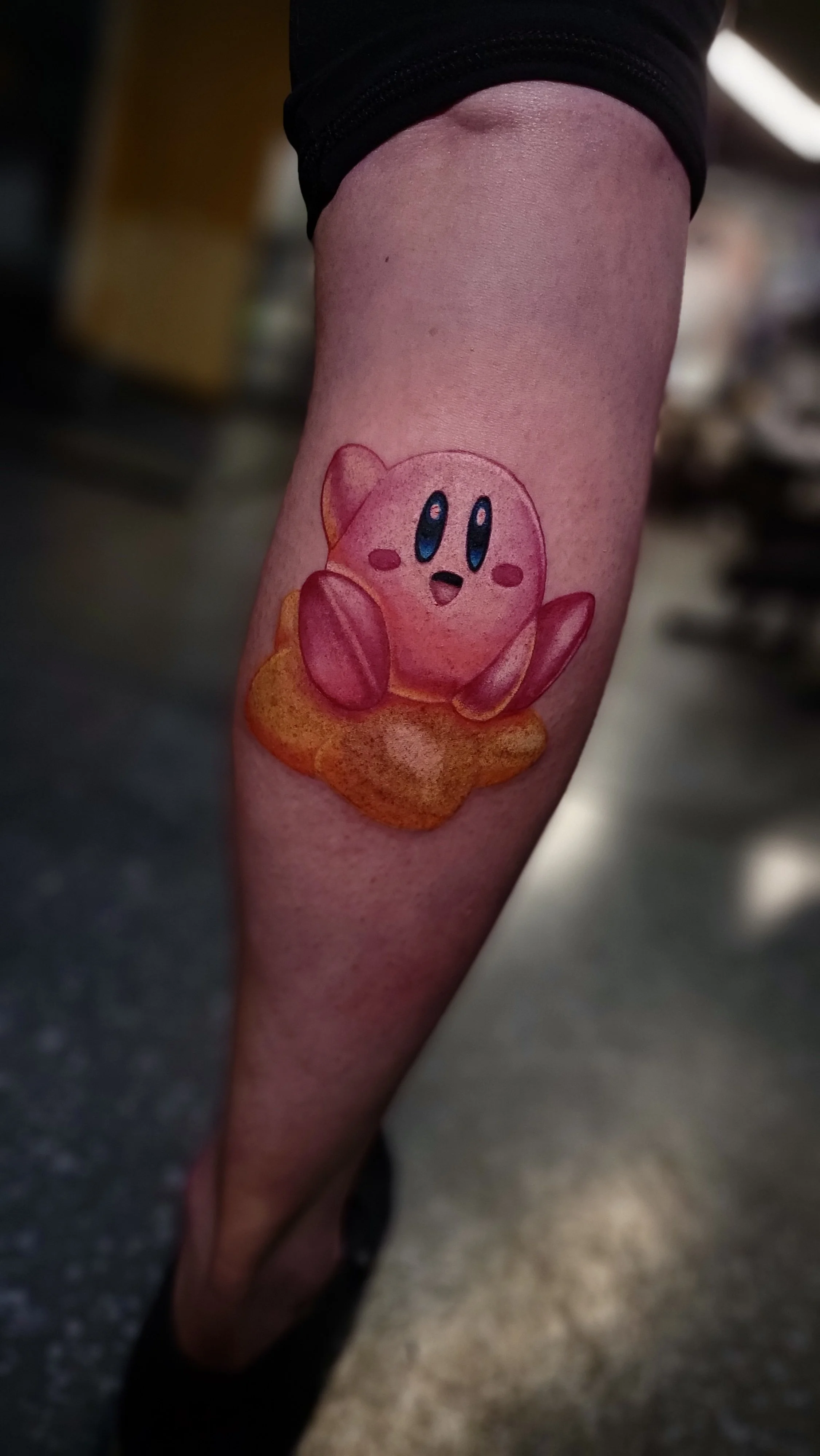 Tattoo of Kirby, the pink video game character, on a person's arm, sitting on a small orange cloud.