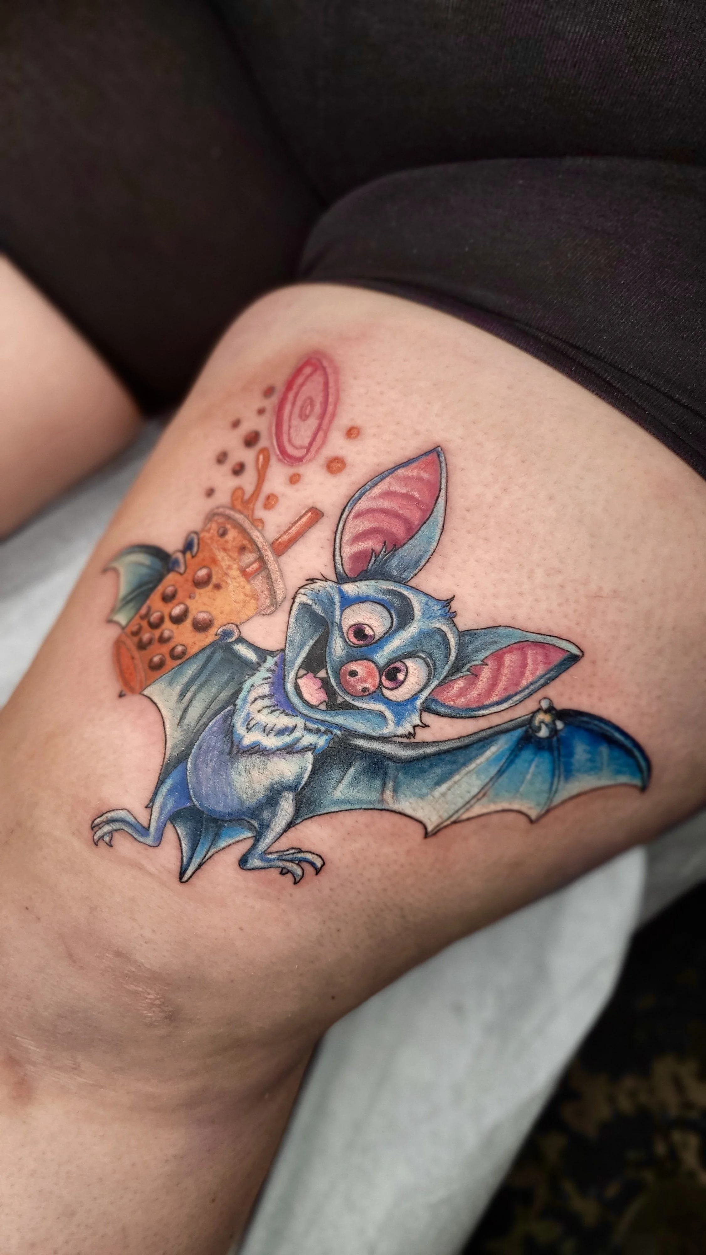 Colorful tattoo of an animated bat with pink ears, big eyes, and a playful expression, holding a bottle of orange soda with a straw.