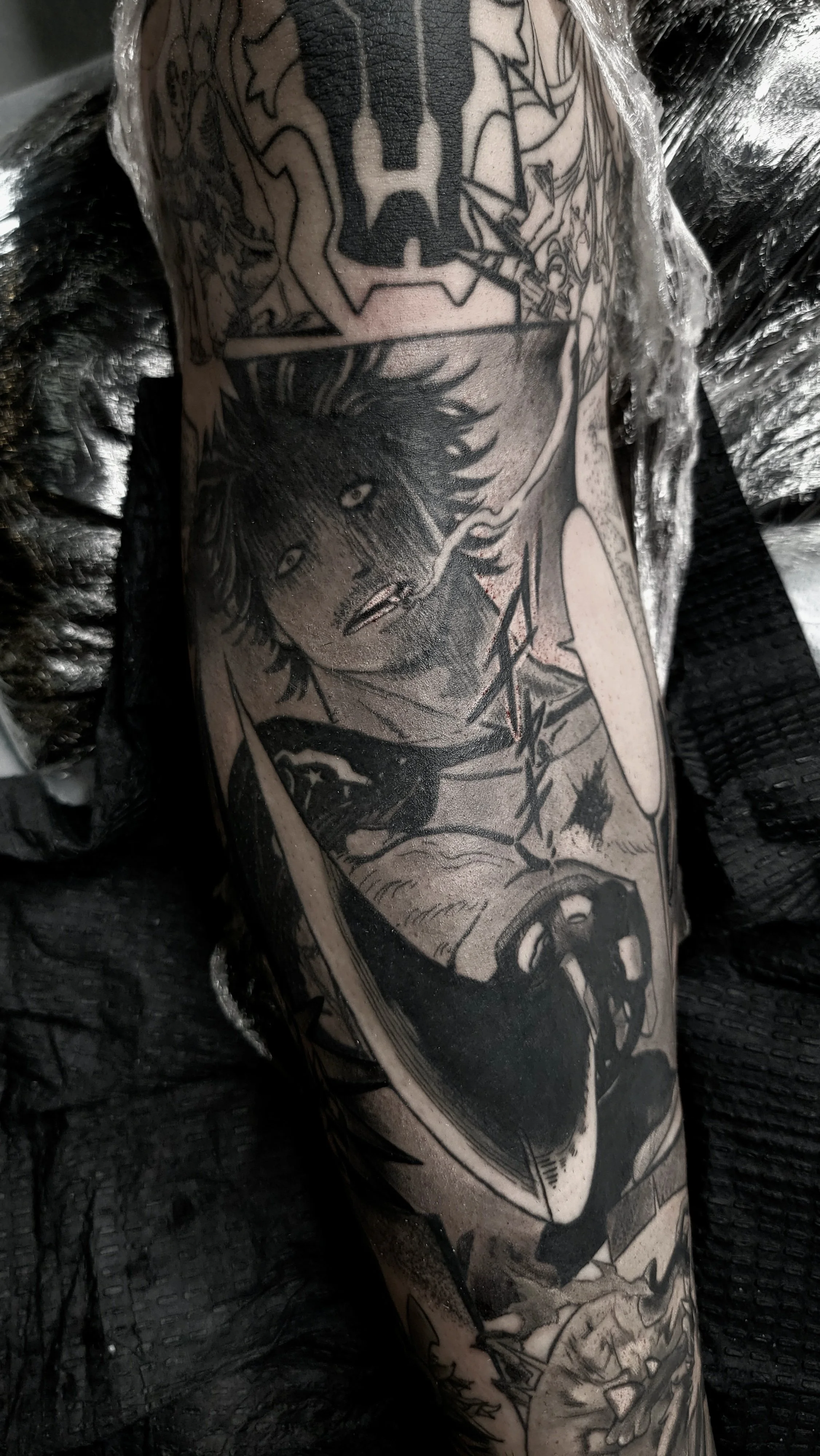 Black and gray tattoo sleeve featuring a human face with wild hair, a shield with a skull, and various abstract designs on a person's arm.