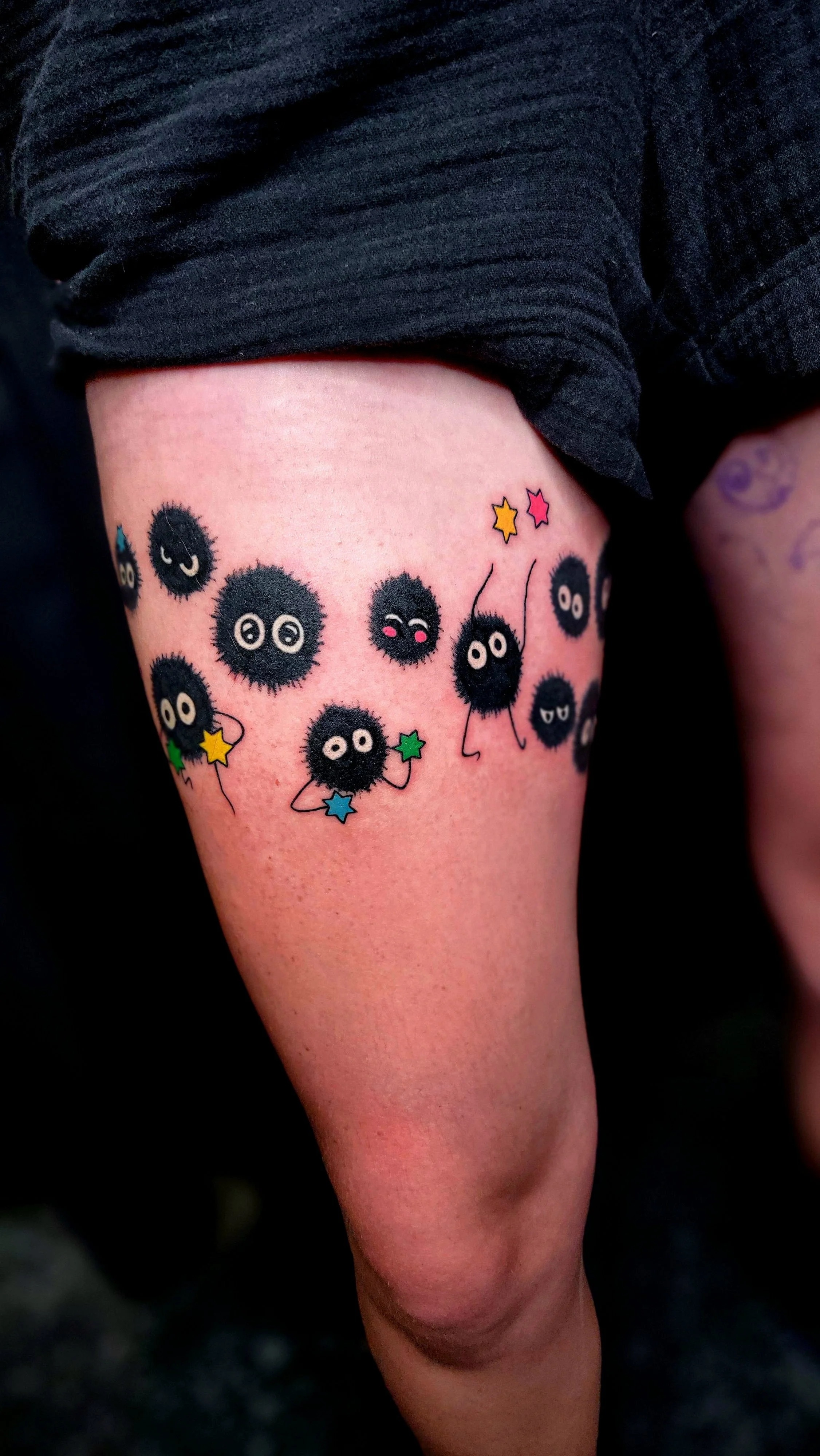 Tattoo of cartoon fluffy black balls with eyes and limbs, some holding colorful stars, on a person's thigh.