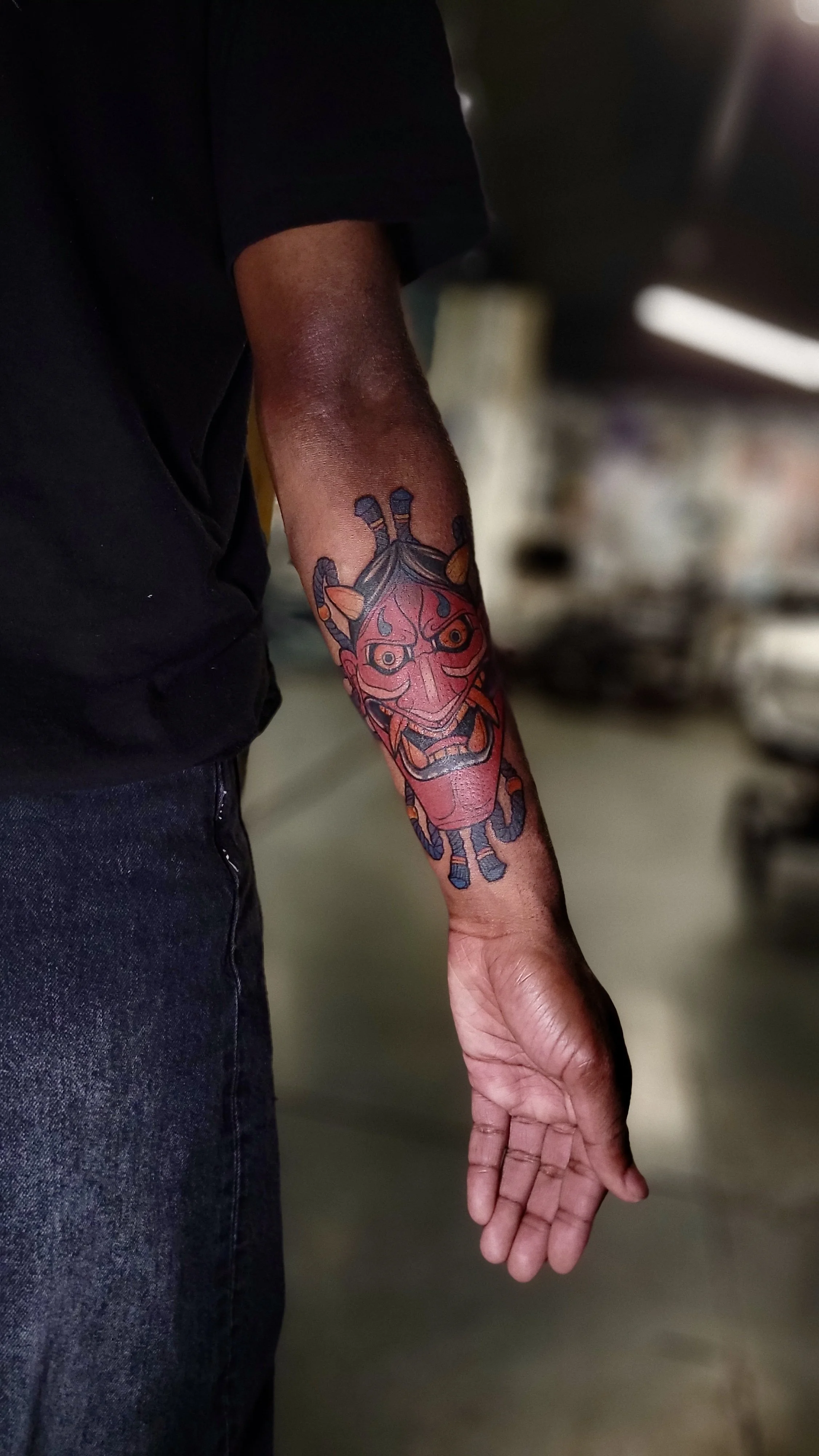 Person with a colorful tattoo of a traditional Asian dragon on their forearm, wearing a black shirt and jeans, standing in a dimly lit indoor space.