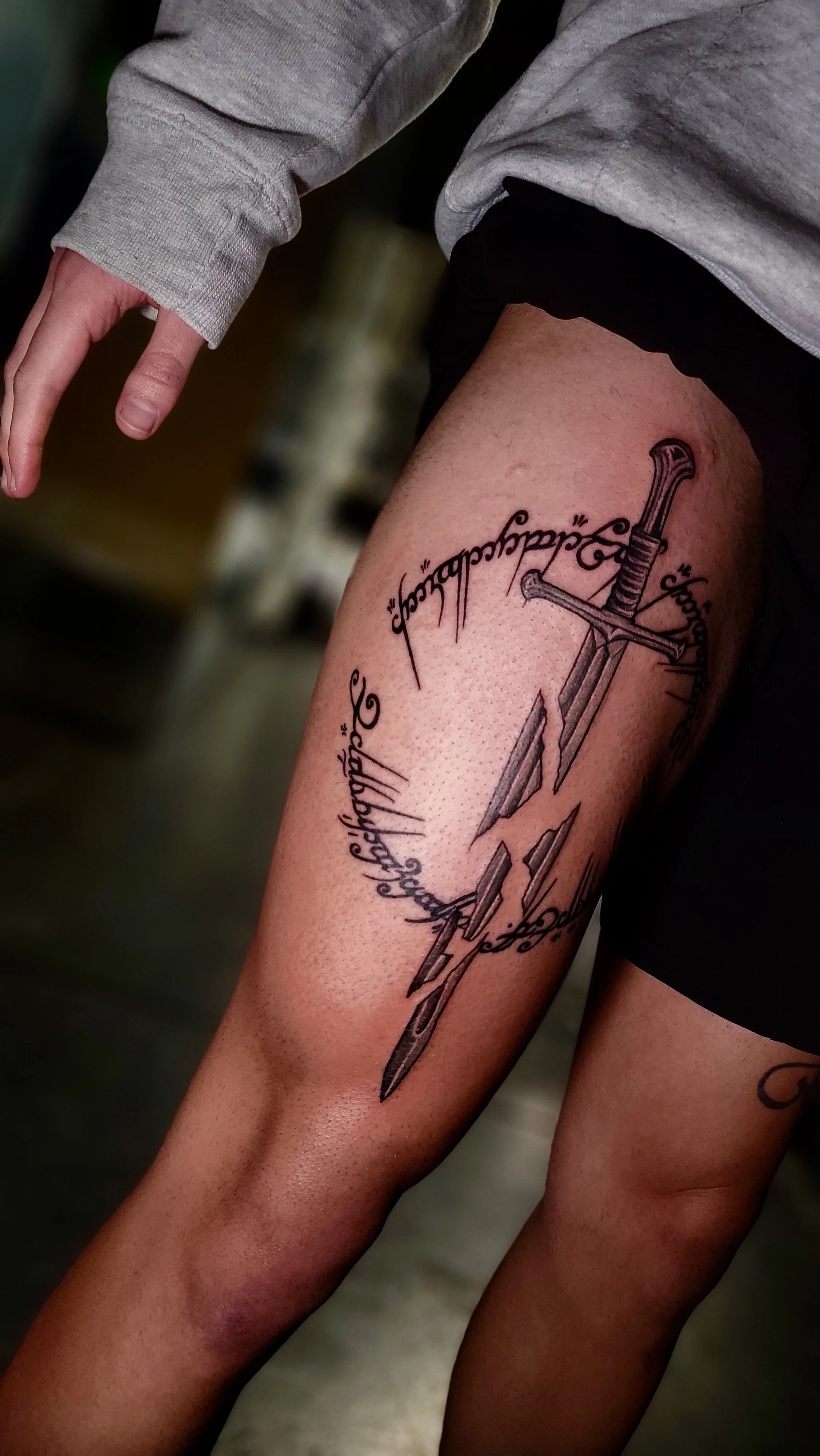 A person's upper thigh with a tattoo of a sword and surrounding text.
