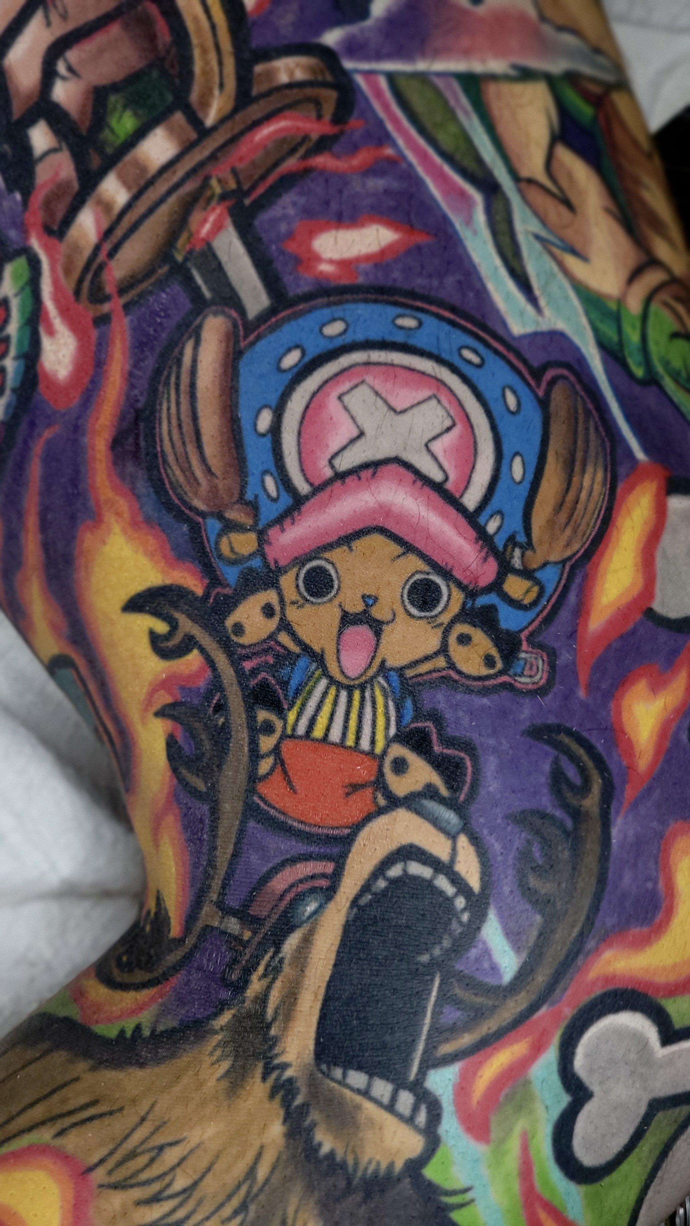 Colorful tattoo featuring a cartoonish animal character with large eyes, a wide-open mouth, and a hat resembling a sheriff's badge. The tattoo includes vibrant colors like purple, red, yellow, blue, green, and orange, with flames and other decorative elements surrounding the character.