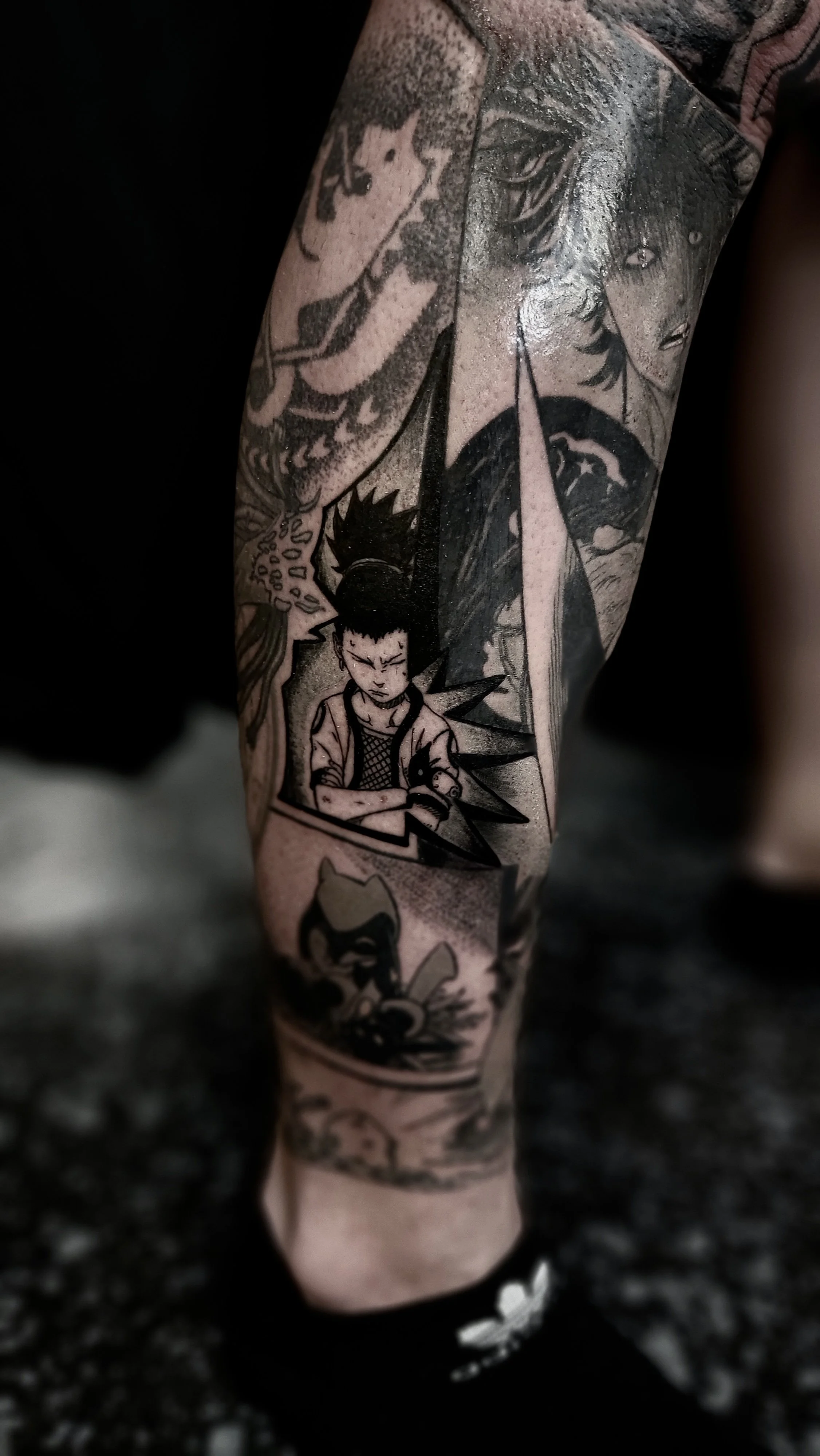 Tattoo sleeve featuring comic book-style characters, including a young boy with spiky hair and a serious expression, and other stylized figures and elements from comic art in black and gray ink.