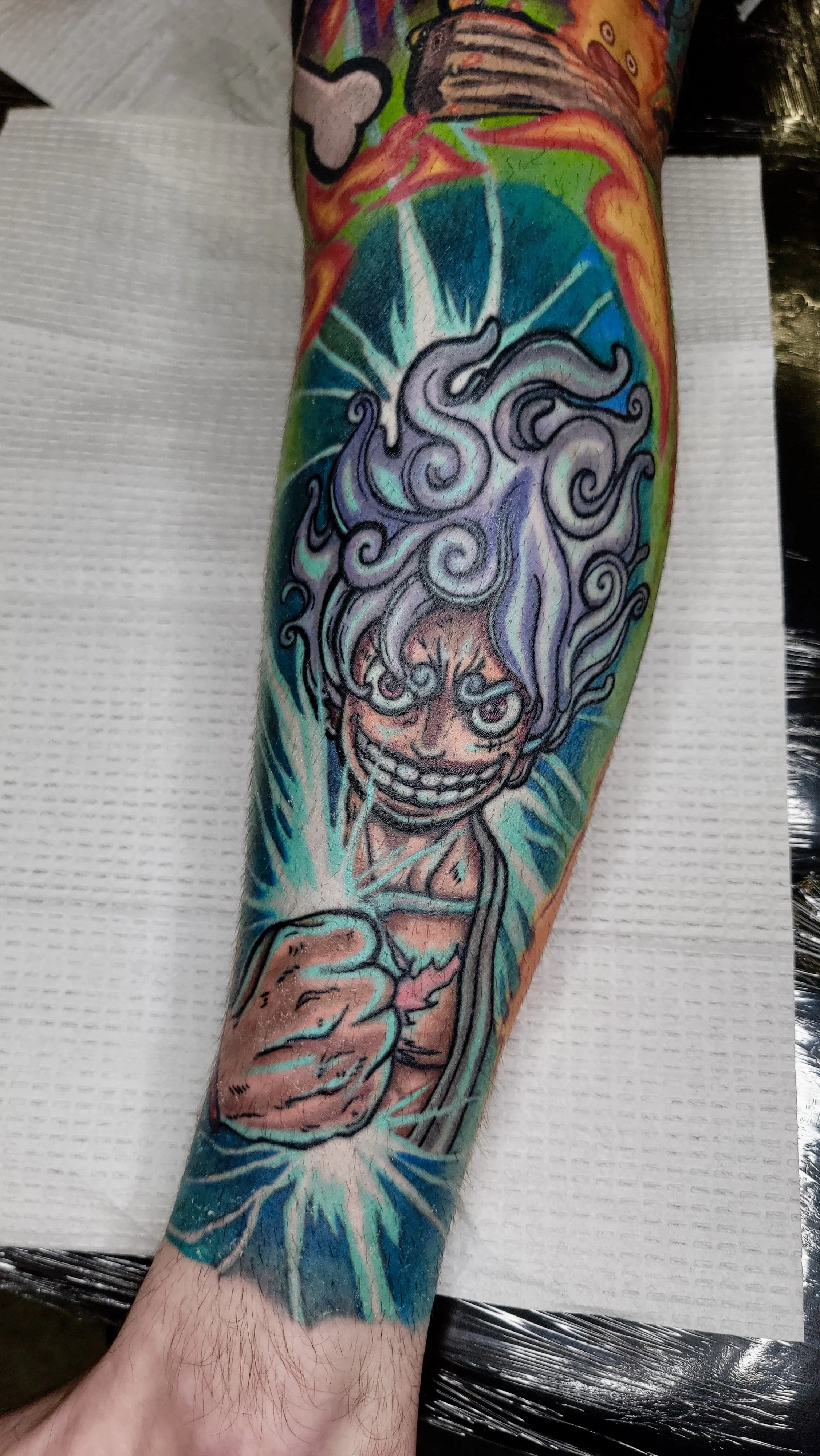 Colorful tattoo on person's arm featuring a grinning skull with swirling smoke, lightning, and vibrant background elements.