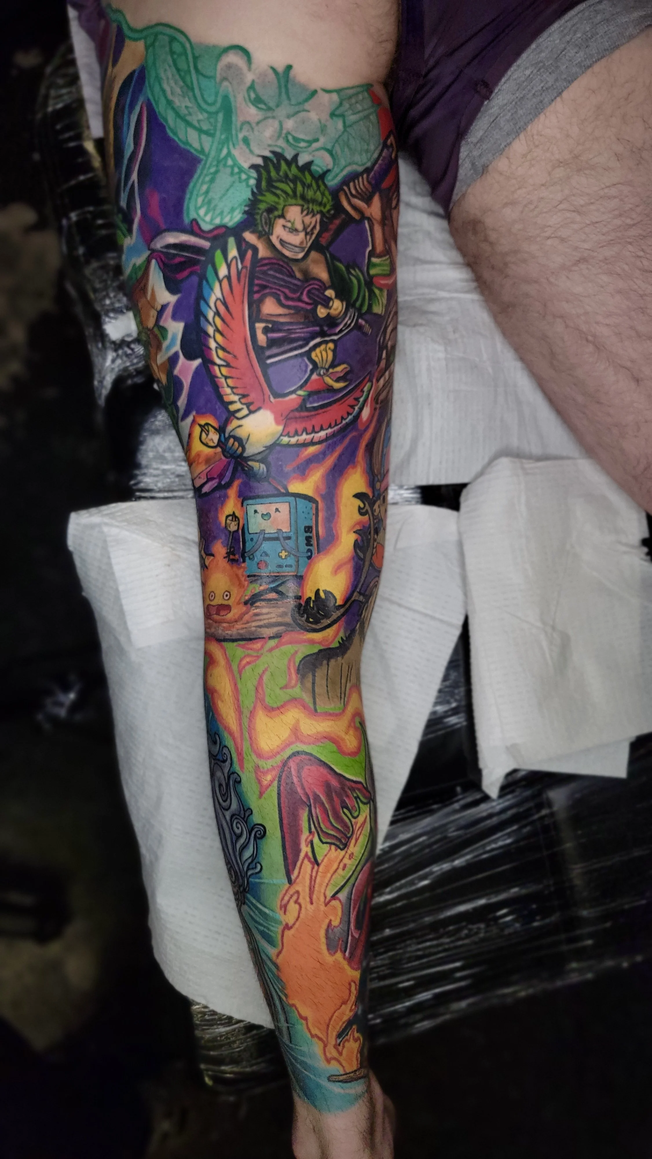 Colorful tattoo sleeve featuring a fierce armored warrior with green hair, a dragon, a bird, a retro game console, and a fiery landscape with swirling flames and abstract patterns.