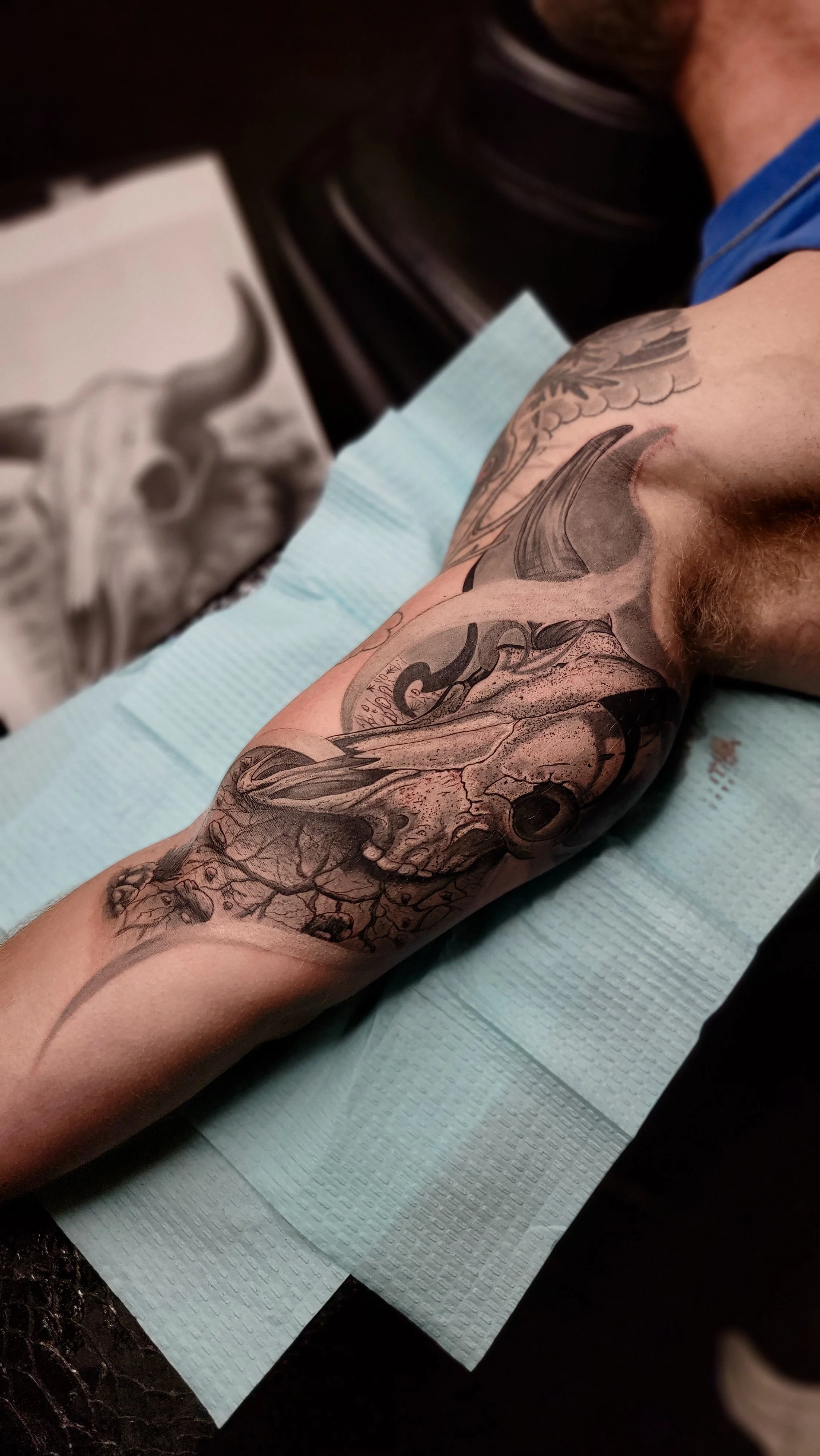 Tattooed arm with detailed skull and floral design resting on a blue medical pad.