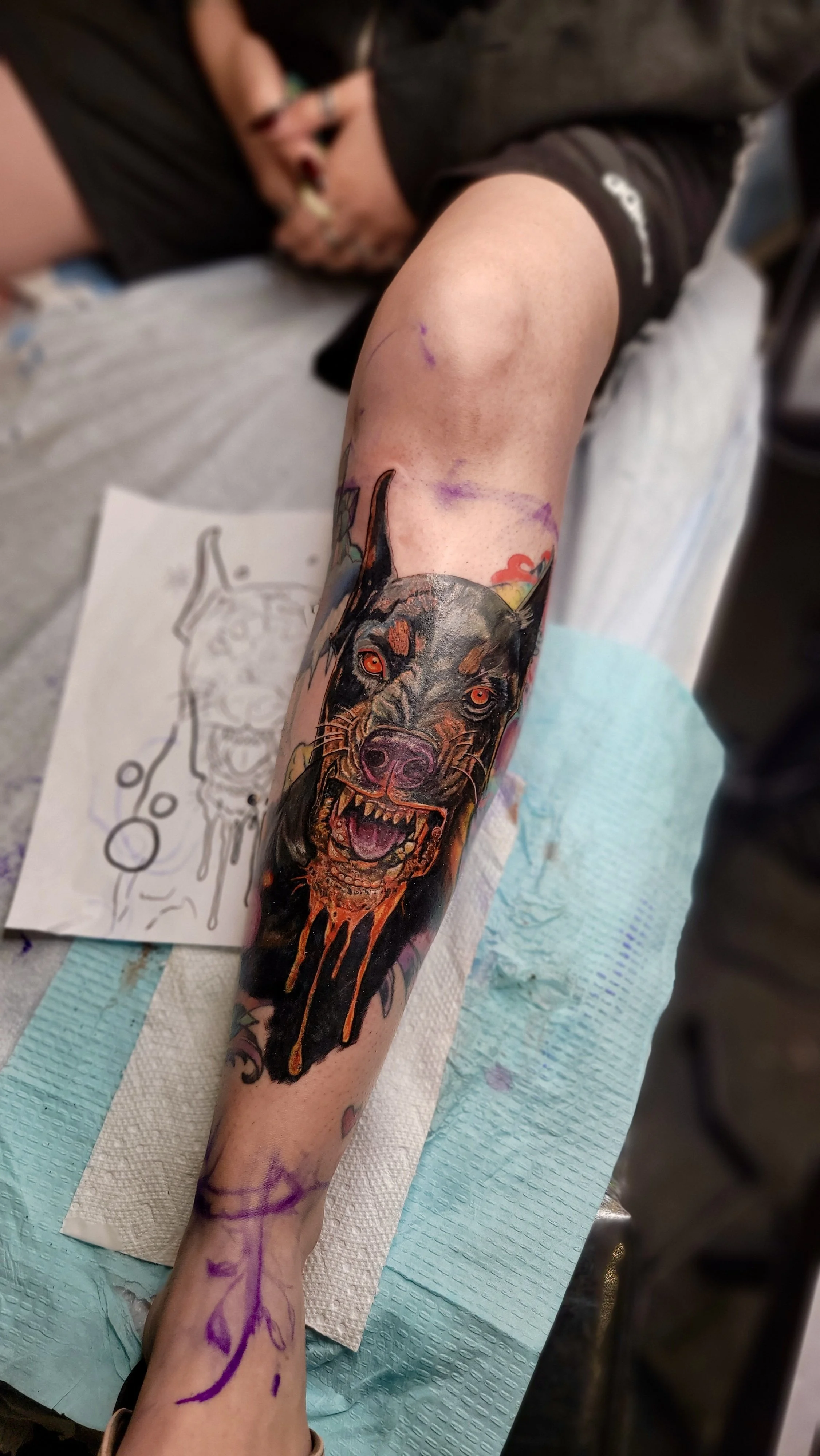 A person is lying on a table with a tattoo of a snarling, aggressive dog with orange eyes and dripping orange and black ink on their forearm. The person is in the process of getting the tattoo, with some purple markings and sketch paper nearby.