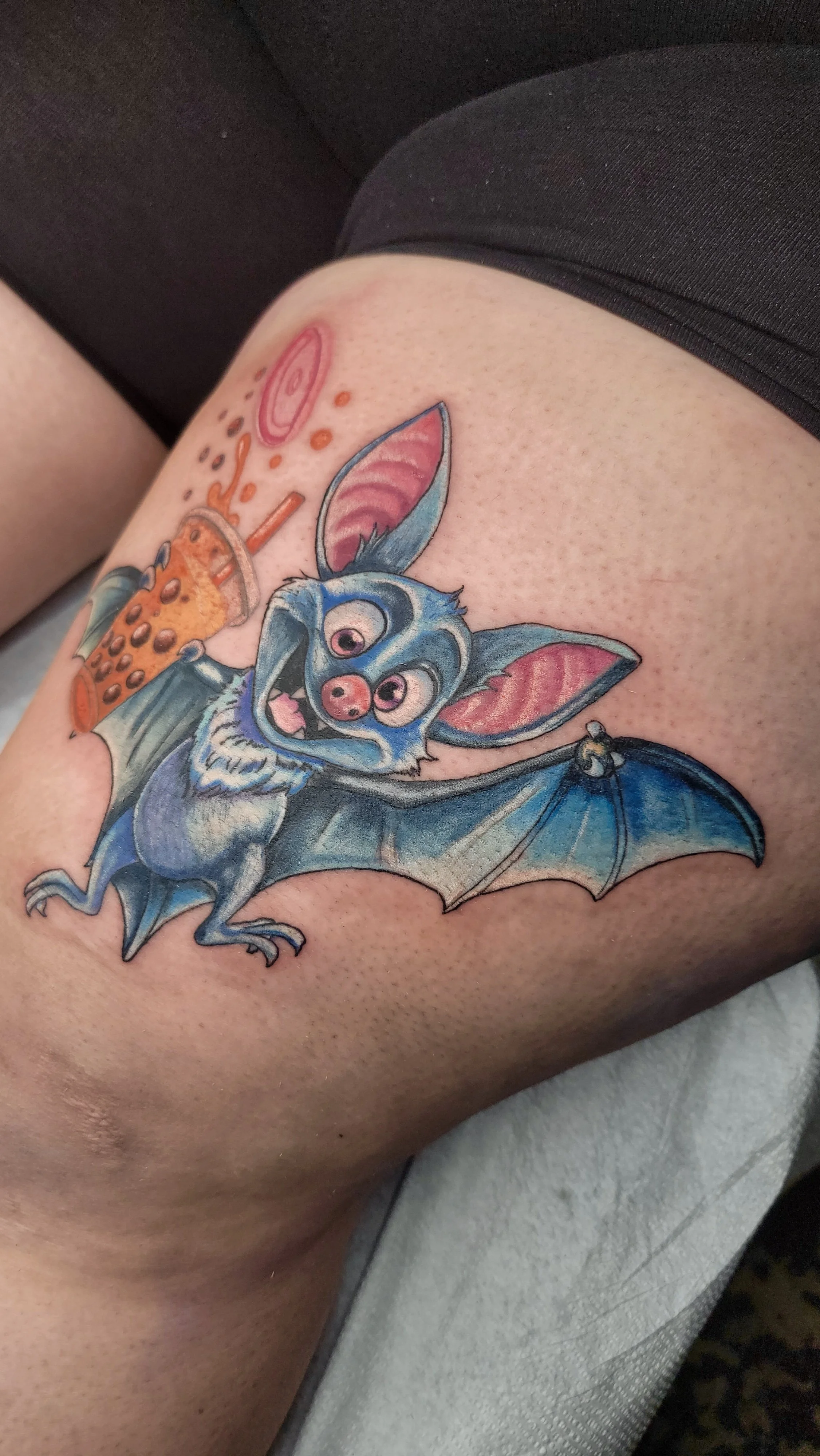 Colorful tattoo of a cartoonish bat with big eyes, pink nose, pink and black ears, blue body, and wings, with a bubble tea drink and colorful toppings.