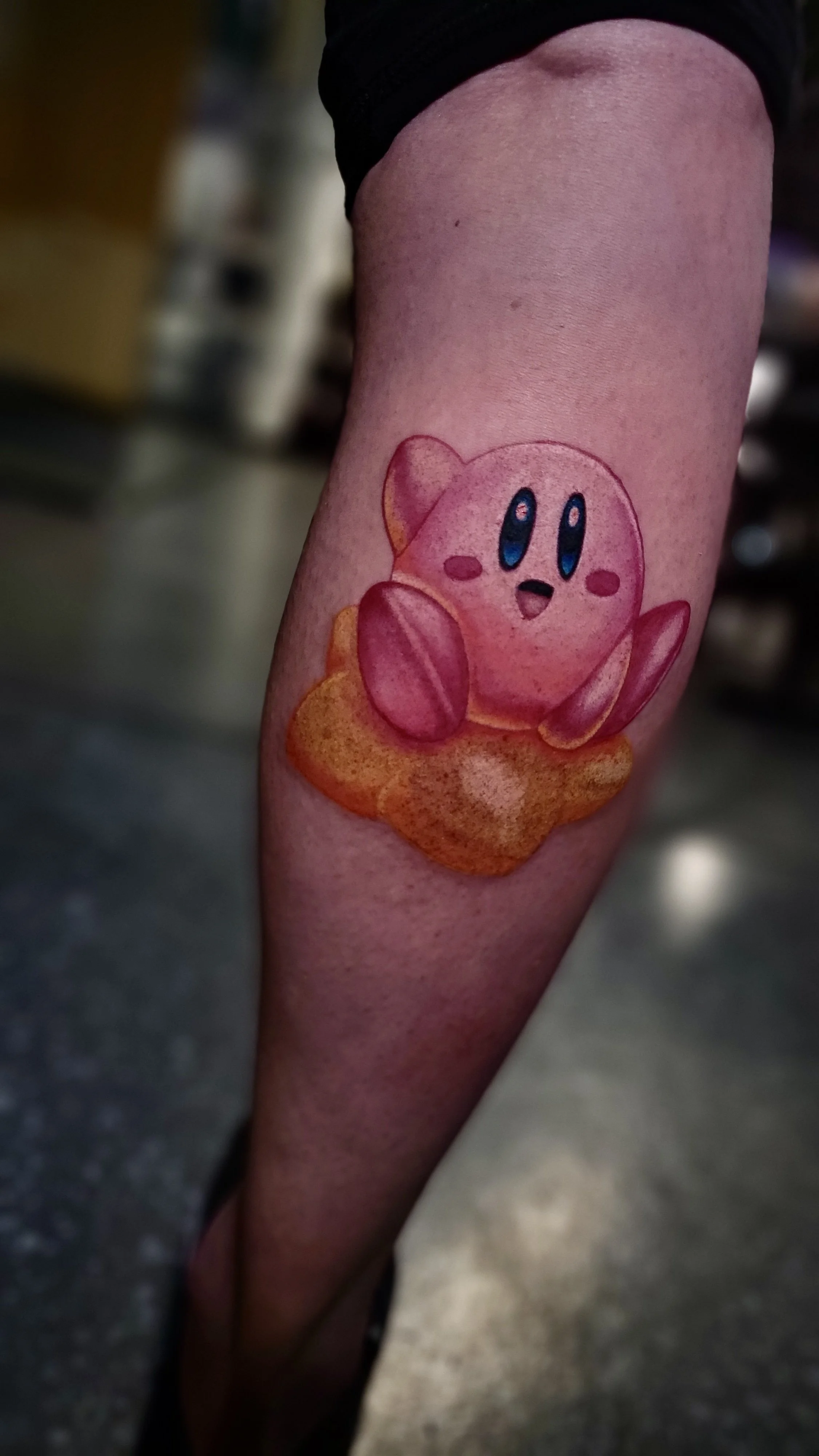 Colorful tattoo of Kirby, a pink animated character with big blue eyes and a cheerful expression, sitting on an orange cloud on a person's arm.