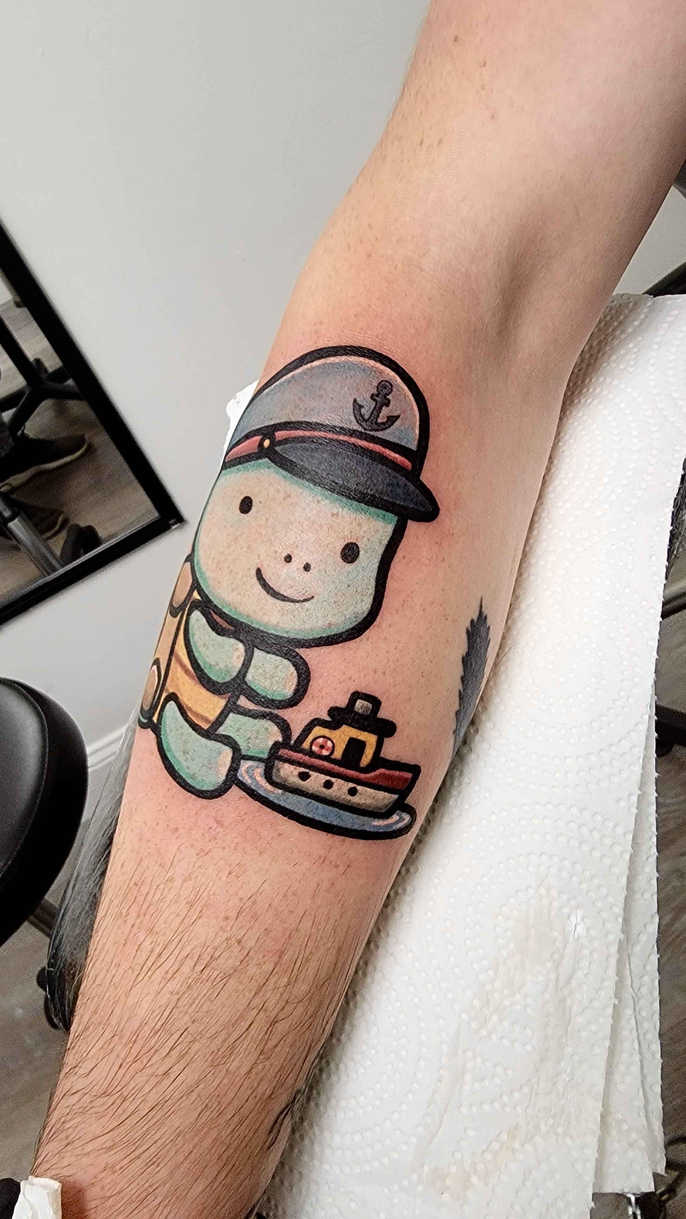 Colorful tattoo of a cute personified submarine with a smiling face, wearing a sailor hat with an anchor, holding a small boat, on a person's arm.
