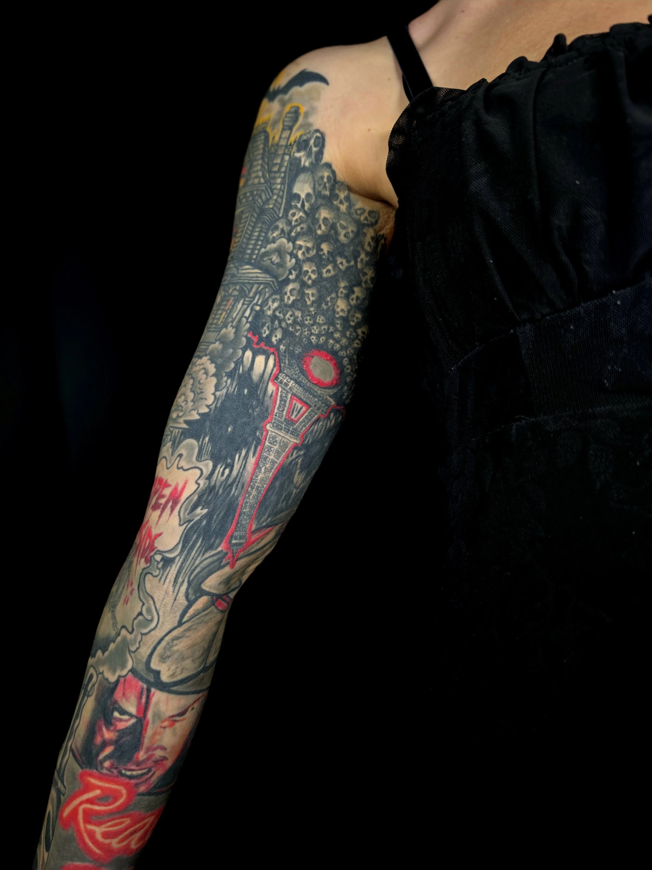 A person's arm with a colorful tattoo sleeve featuring a cityscape, skulls, clouds, and a large red and black sword against a black background.