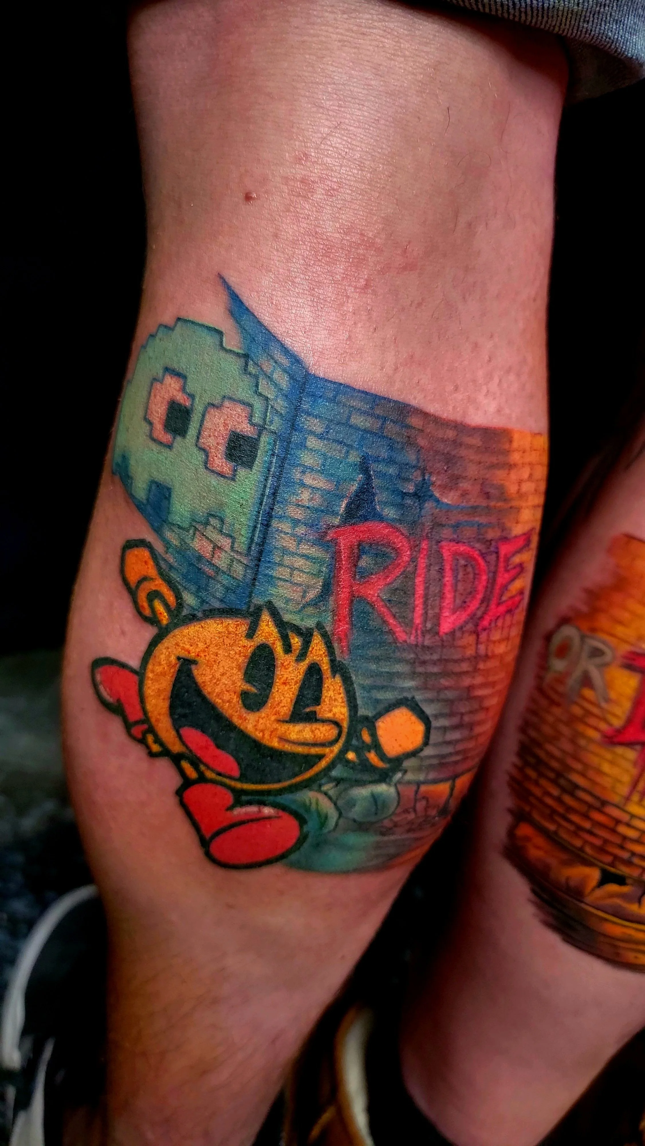 Colorful tattoo of a pixelated skull with the words 'RIDE' next to a city skyline and a pumpkin character with a smiley face, on a person's arm.