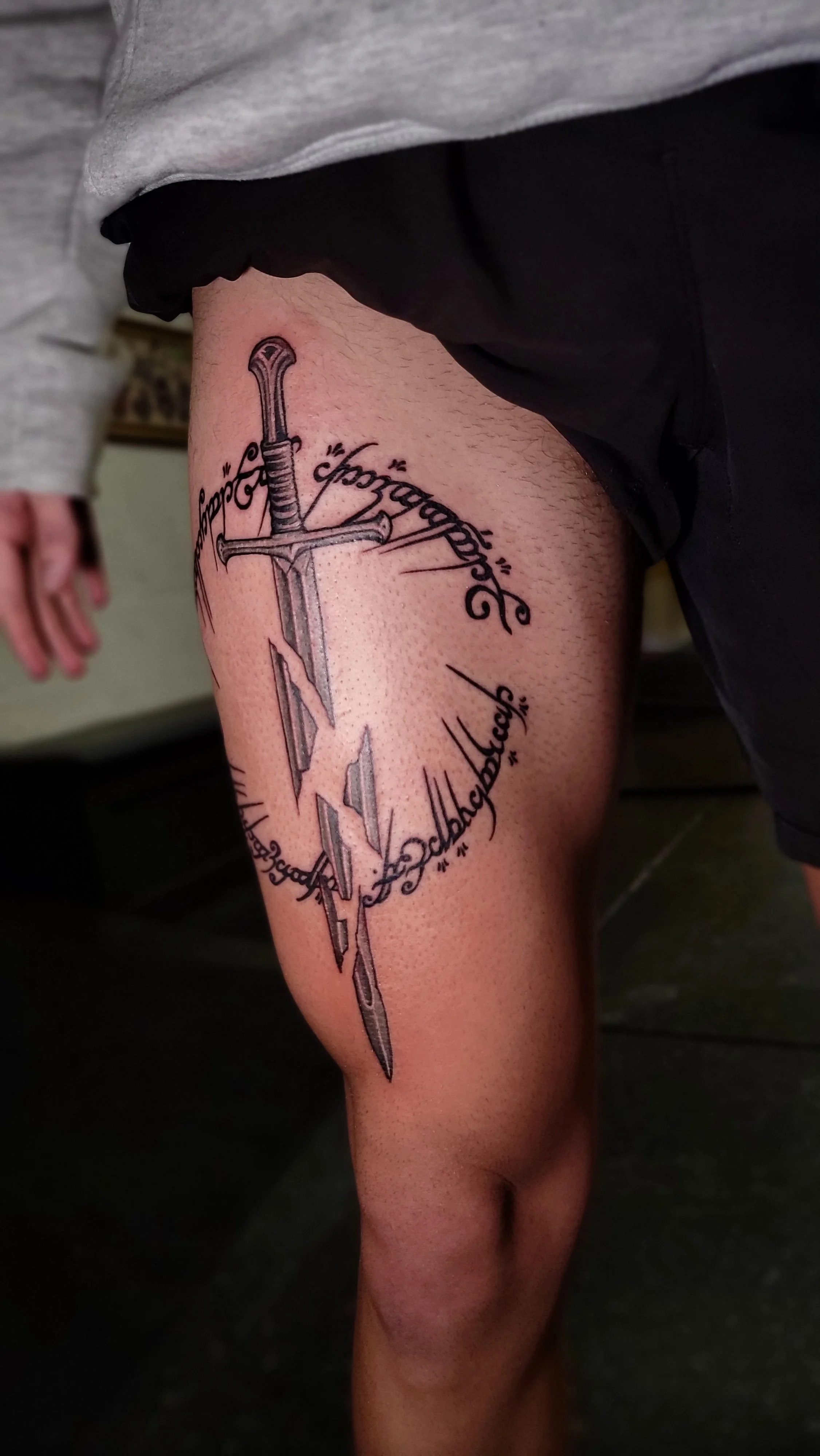Tattoo of a sword with a quote wrapped around the blade on a person's thigh.