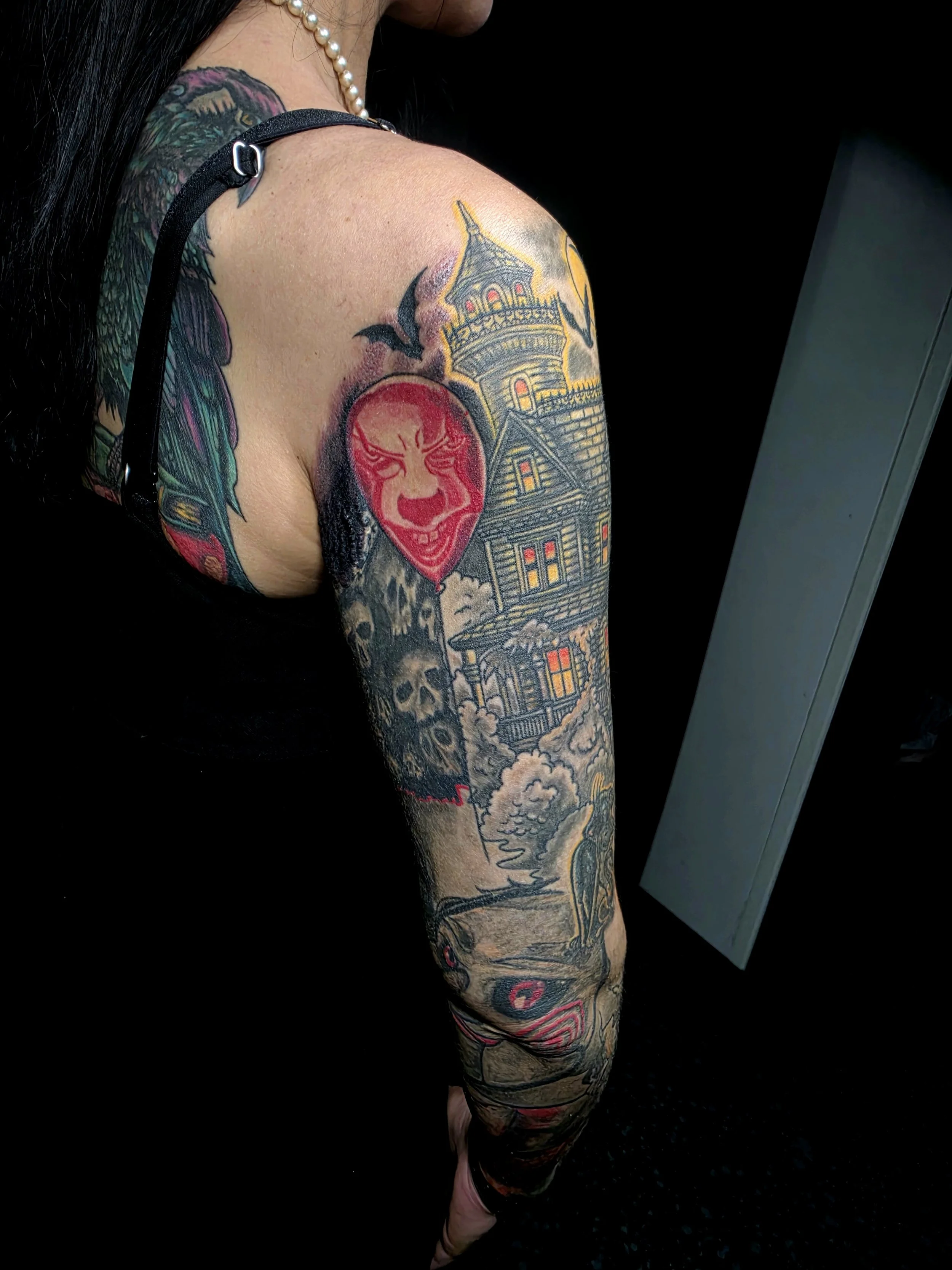 A person's arm with multiple colorful tattoos, including a house with a tower, a clown face, a skull, and various designs, against a black background.