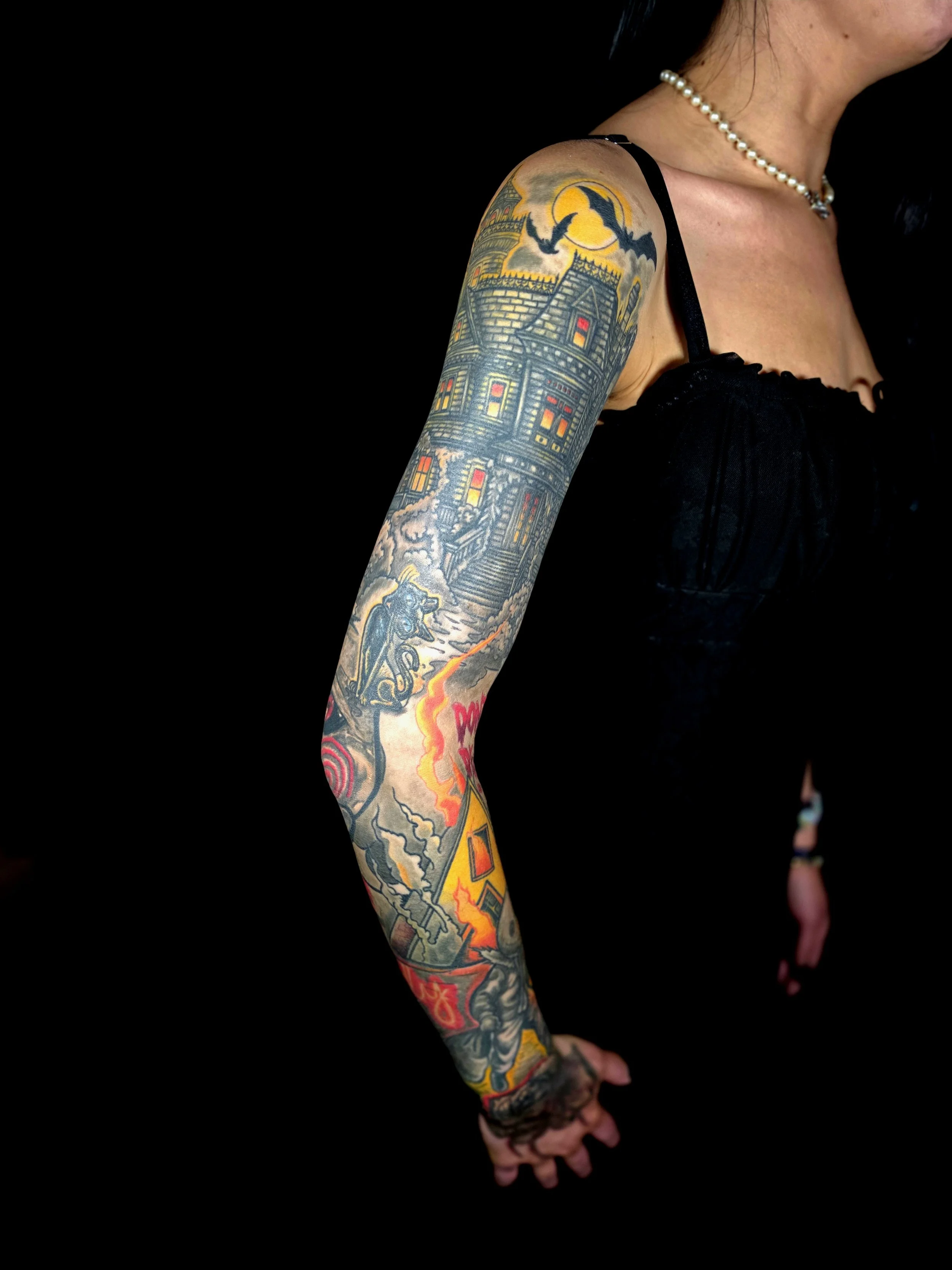 A person's arm covered in colorful tattoos, including a haunted house, bats, a witch on a broomstick, and a lightning bolt, with a dark background.