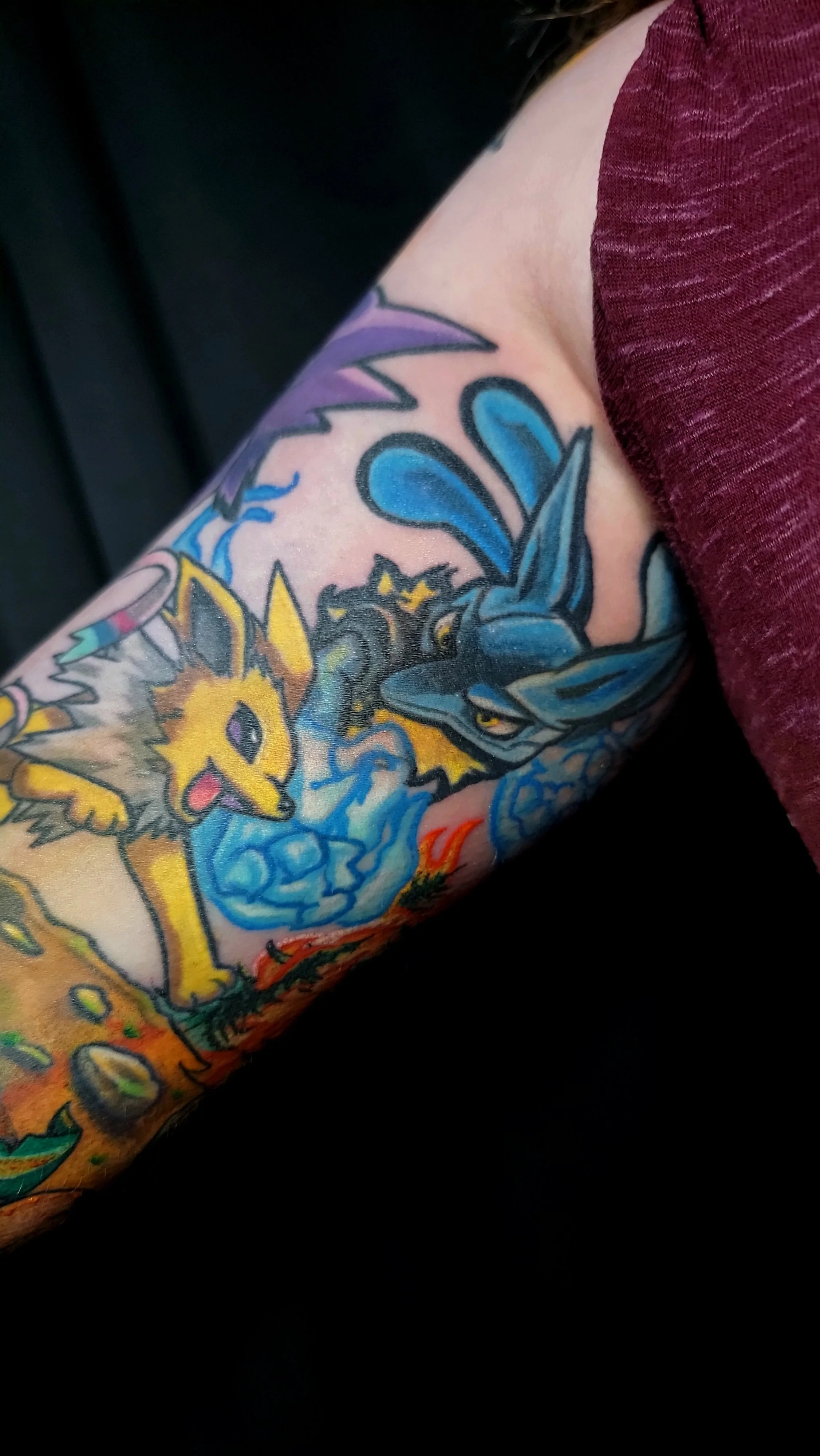 Close-up of a colorful tattoo on a person's arm, featuring Pokémon characters Pikachu and Lucario surrounded by vibrant colors and patterns.